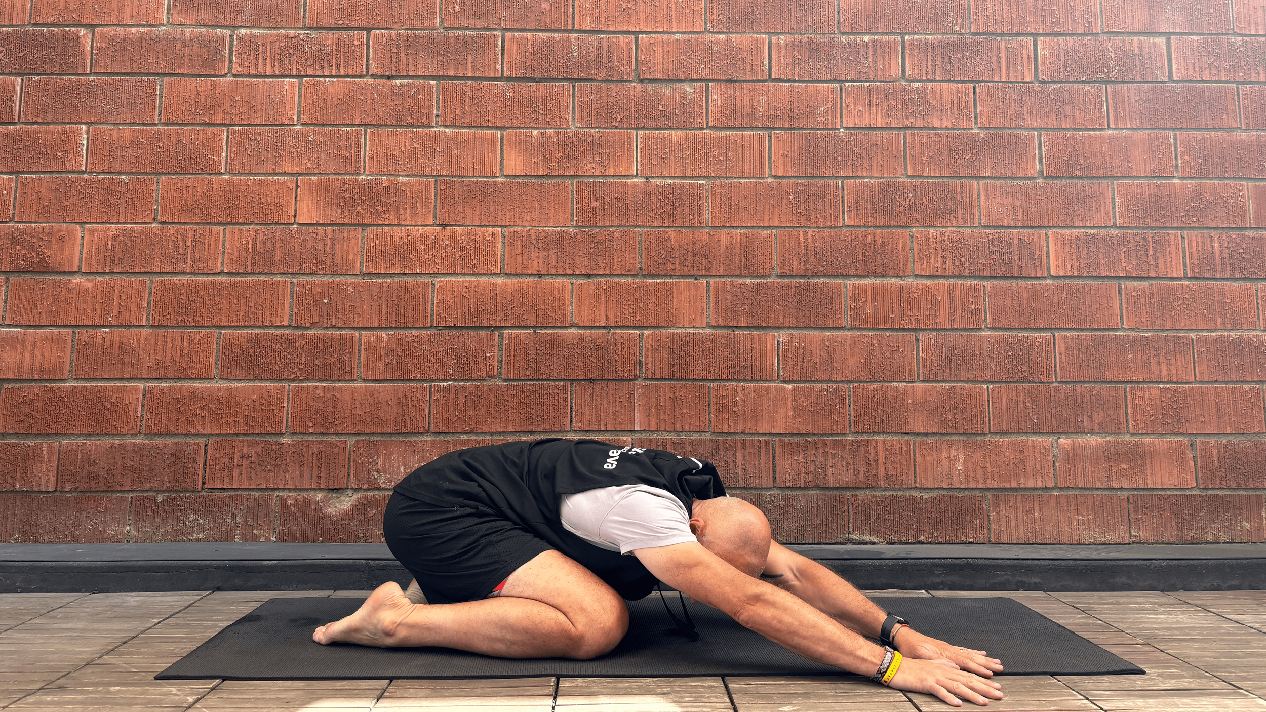young man kneeling on a black yoga mat on the floor with brick wall in the background, stretching arms forward, walking his hands to the right to deepen the stretching on his back