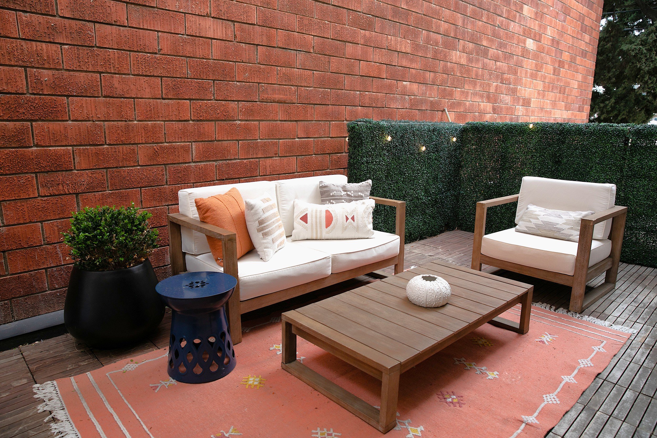 An outdoor patio with a white cushioned sofa and matching armchair, wooden coffee table, coral rug, and decorative pillows, set against a red brick wall and green hedge.