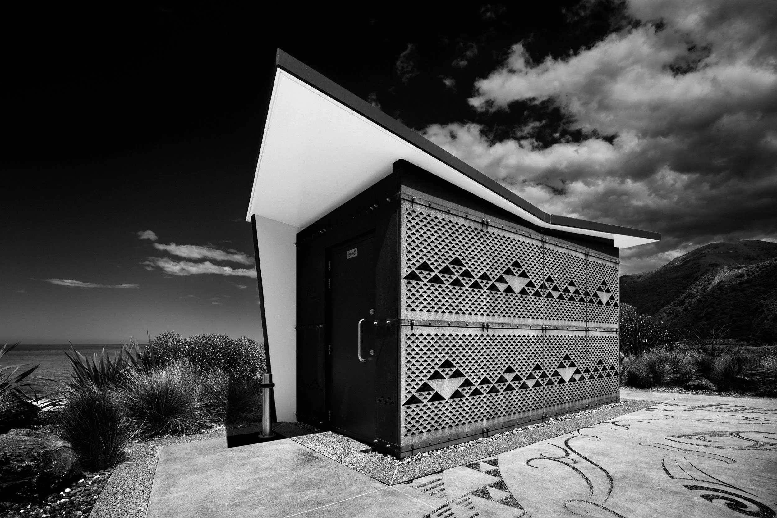 A modern small building with a geometric patterned facade, set in a landscaped area with bushes and a mountain background, under a cloudy sky, in black and white.