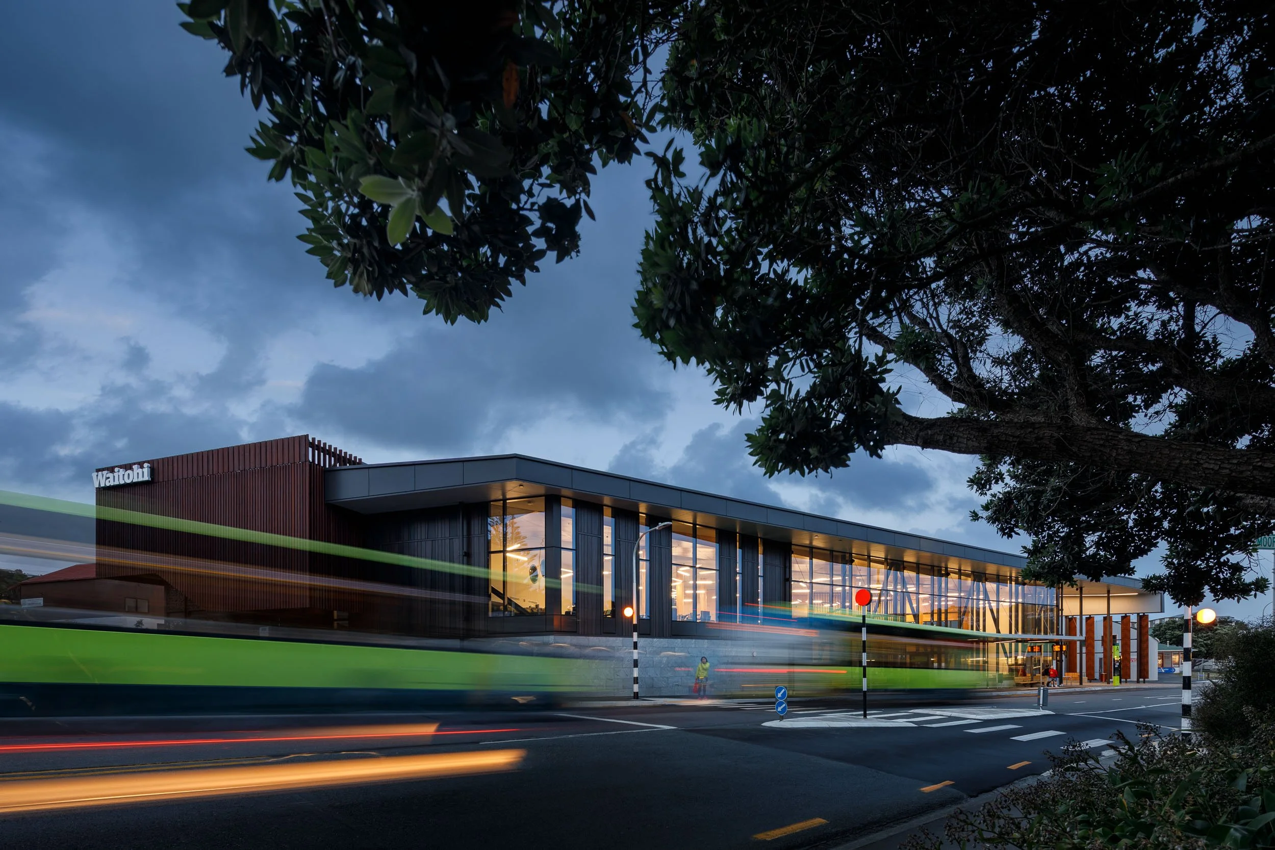 A modern building with large glass windows at dusk, with streaks of light from passing vehicles on the road in front, surrounded by trees and a cloudy sky.