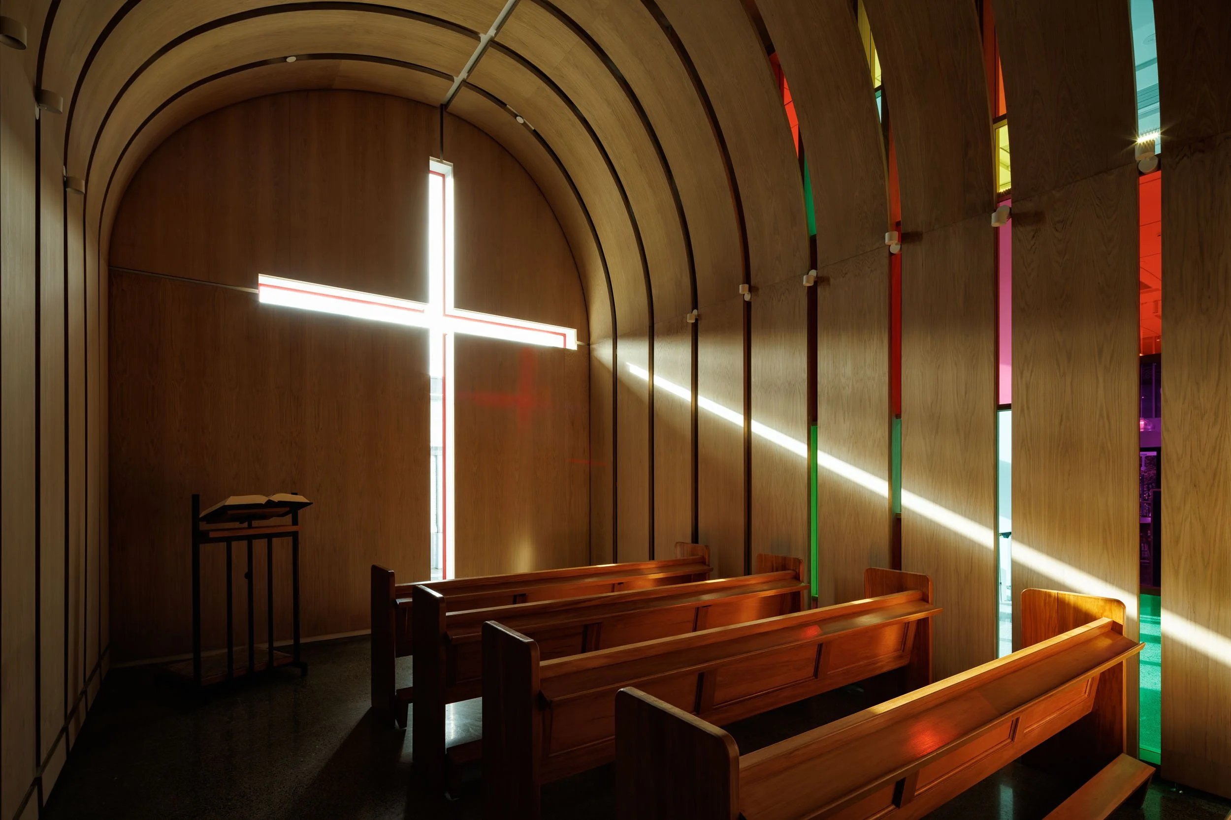 Interior of a small chapel with wooden pews, a cross-shaped window with colorful stained glass, and simple wooden walls.