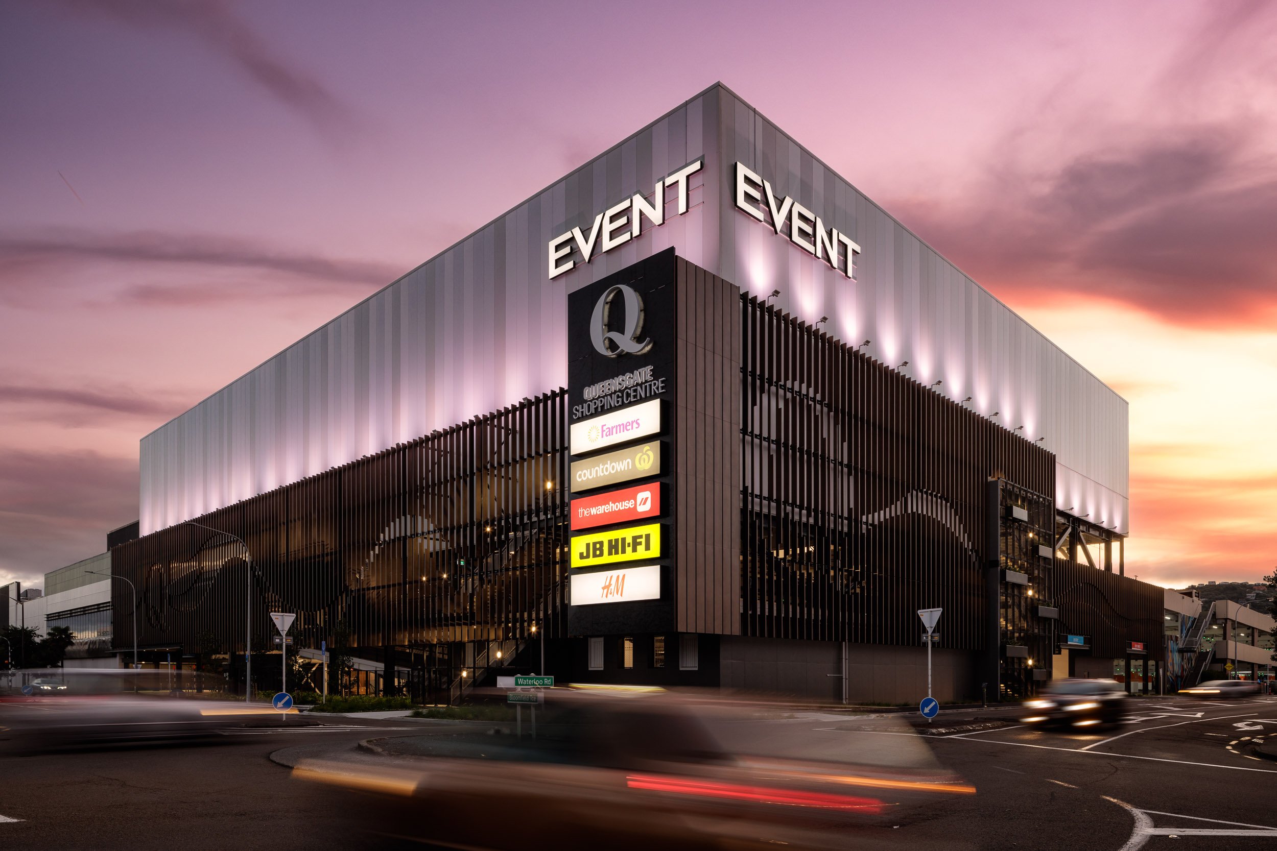 A modern shopping center building with illuminated signs, including brands like Farmers, Countdown, The Warehouse, JB Hi-Fi, and H&M, at sunset with a purple and orange sky.