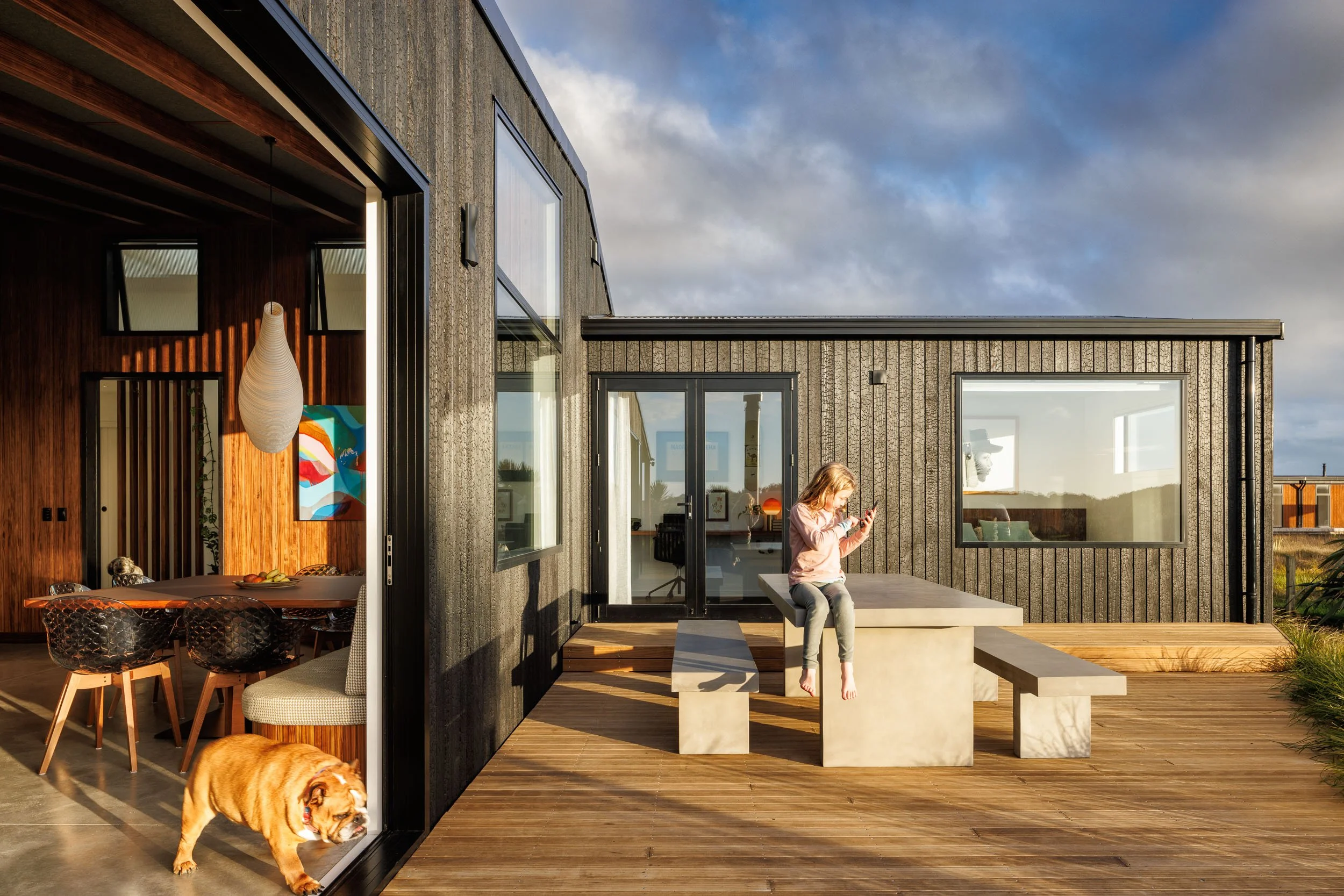A girl sitting on a concrete outdoor table on a wooden deck, looking at her phone. A dog is walking inside through an open sliding glass door, and the interior of a modern home with dining area is visible.