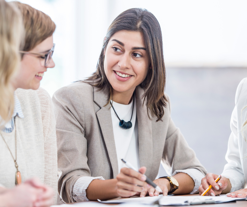 Group of people in a meeting, discussing and smiling, woman in a blazer as the focal point.