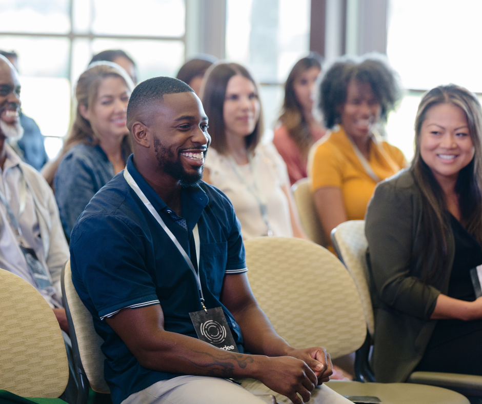 Group of people smiling and listening at an indoor event, wearing conference badges.