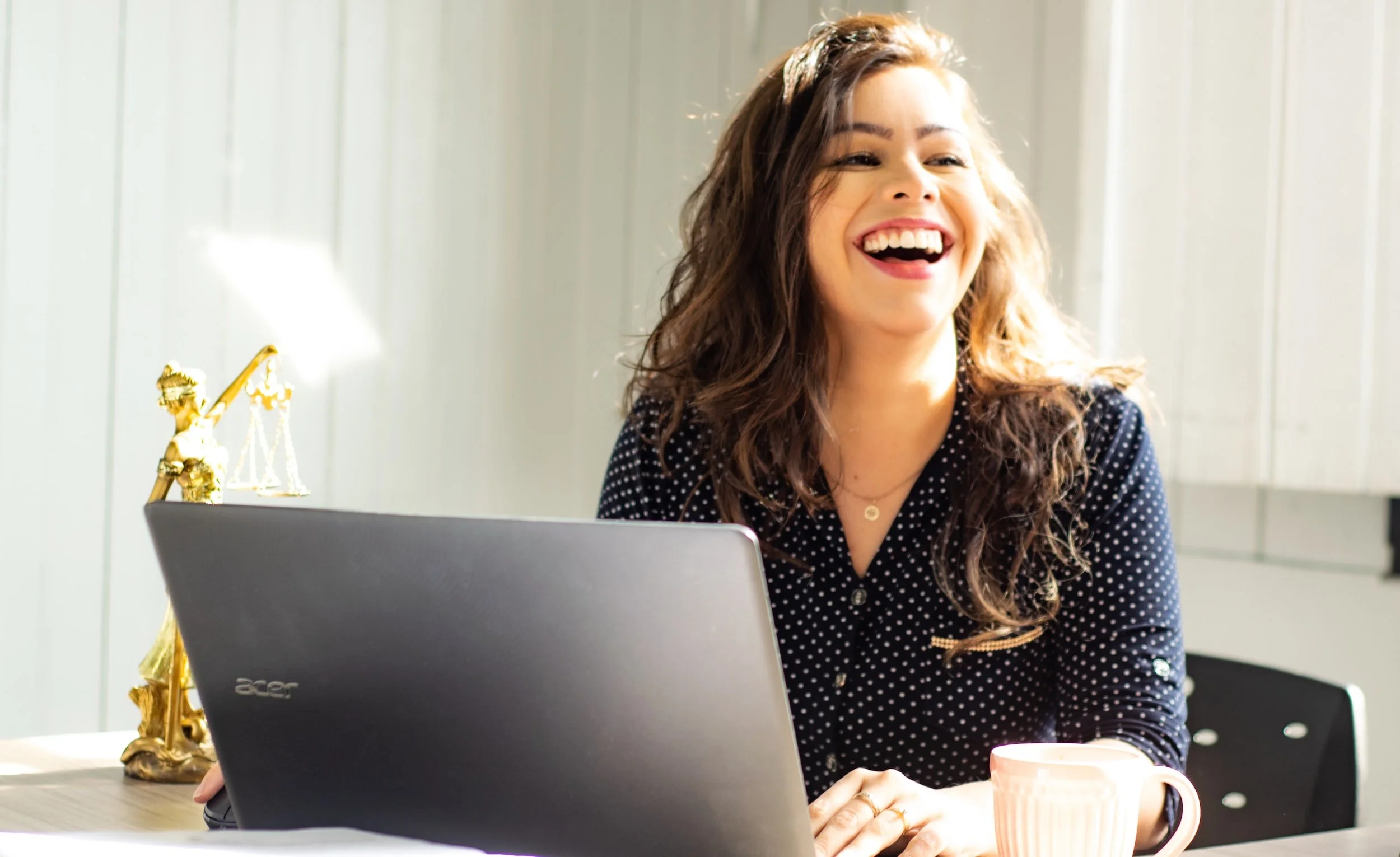 Woman smiling at a desk with a laptop, a cup, and a small statue of Lady Justice nearby.