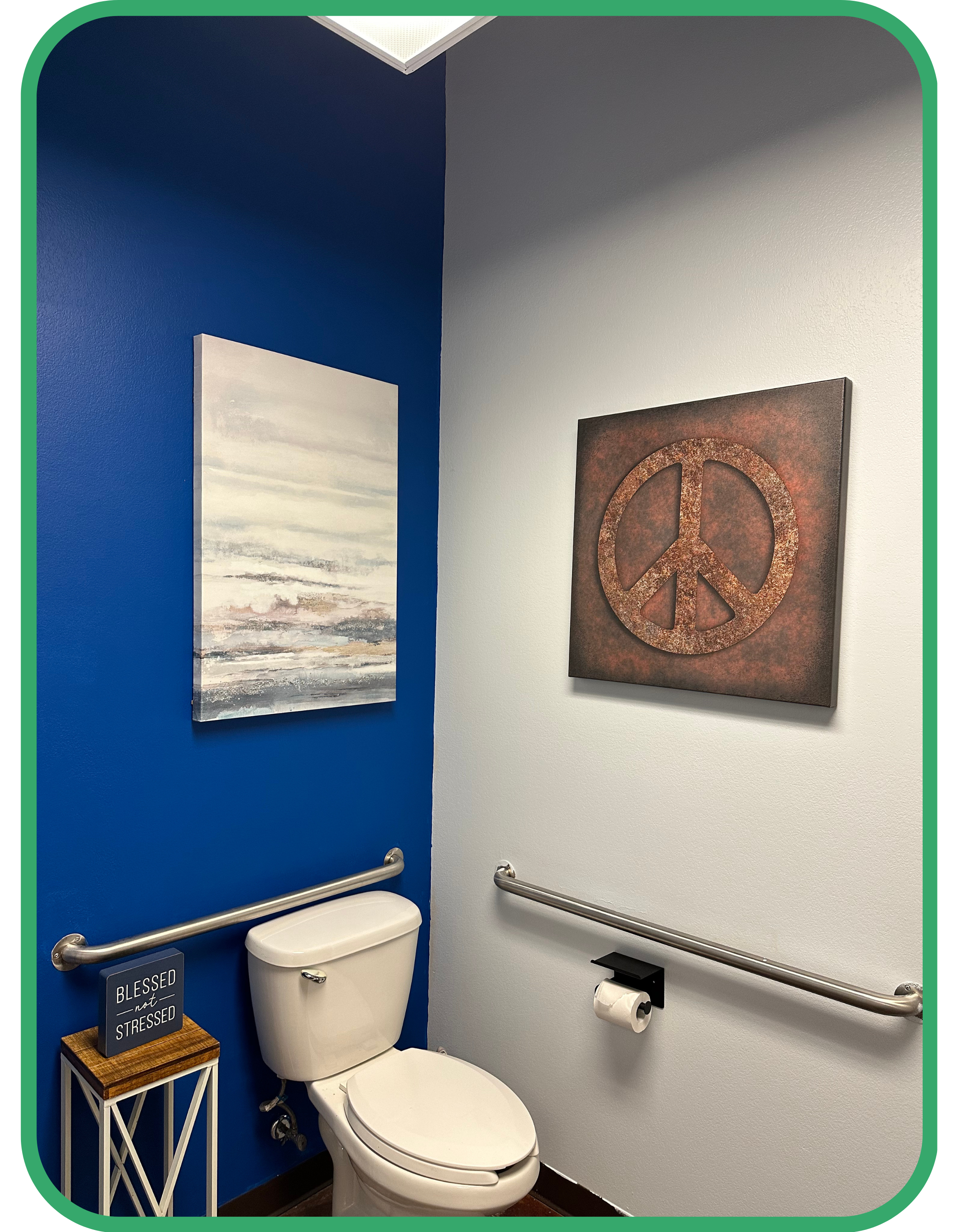 Restroom with blue and white walls, featuring a toilet and safety rails. Wall art includes a peace sign and abstract painting. A small sign on a stand reads 'Blessed not Stressed.'"