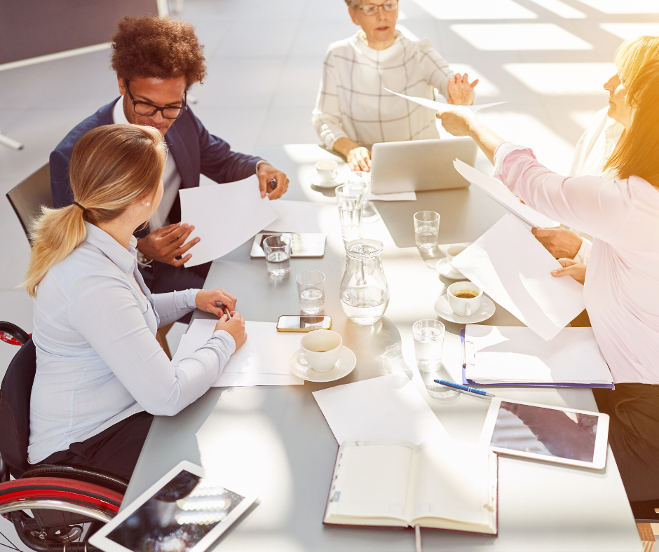 A diverse group of professionals in a modern office setting engaged in a meeting around a table cluttered with documents, coffee cups, and digital tablets.