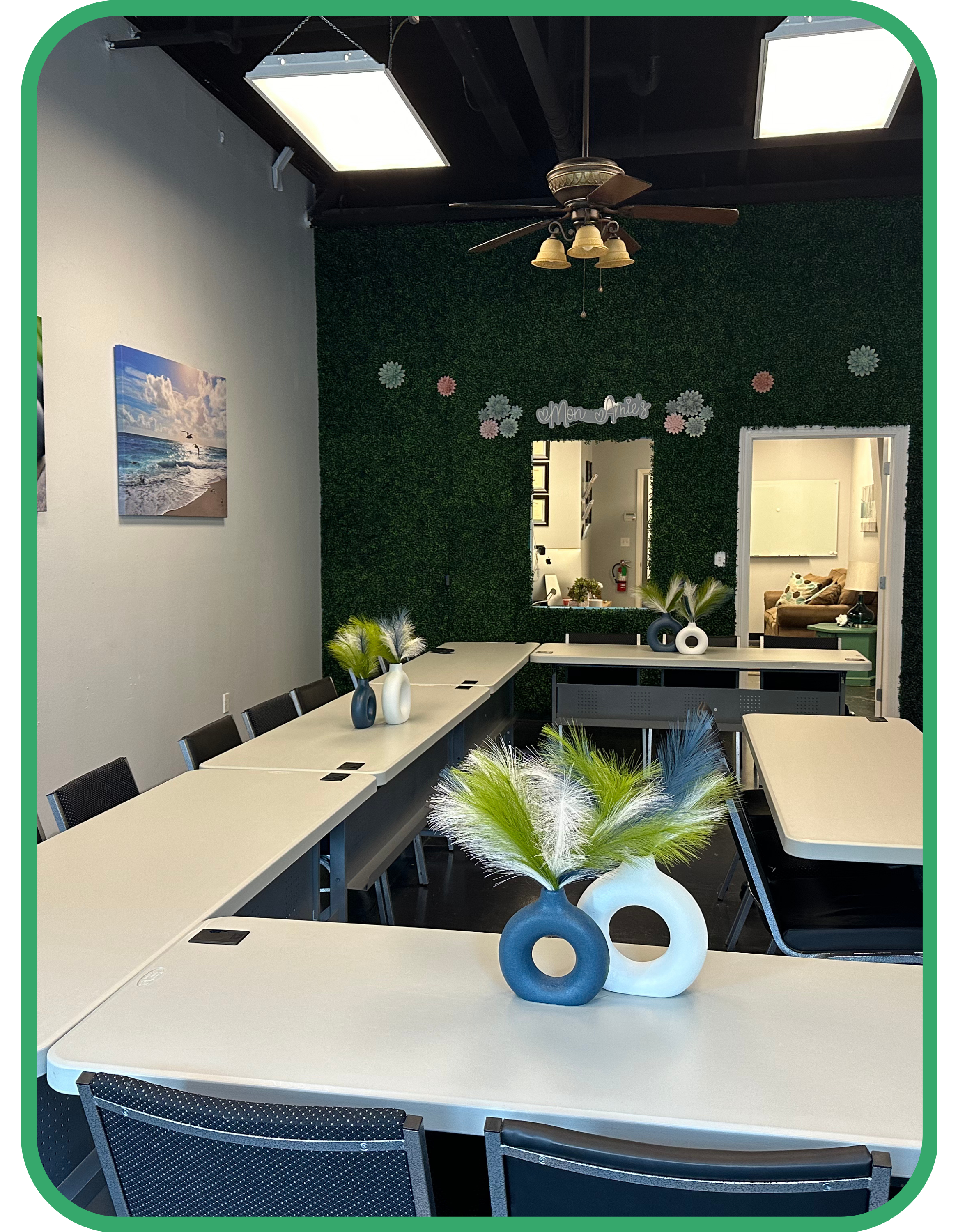 A modern office meeting room with U-shaped tables, decorative vases with greenery, a green accent wall with artificial foliage, and a ceiling fan. The room has framed artwork and is well-lit.