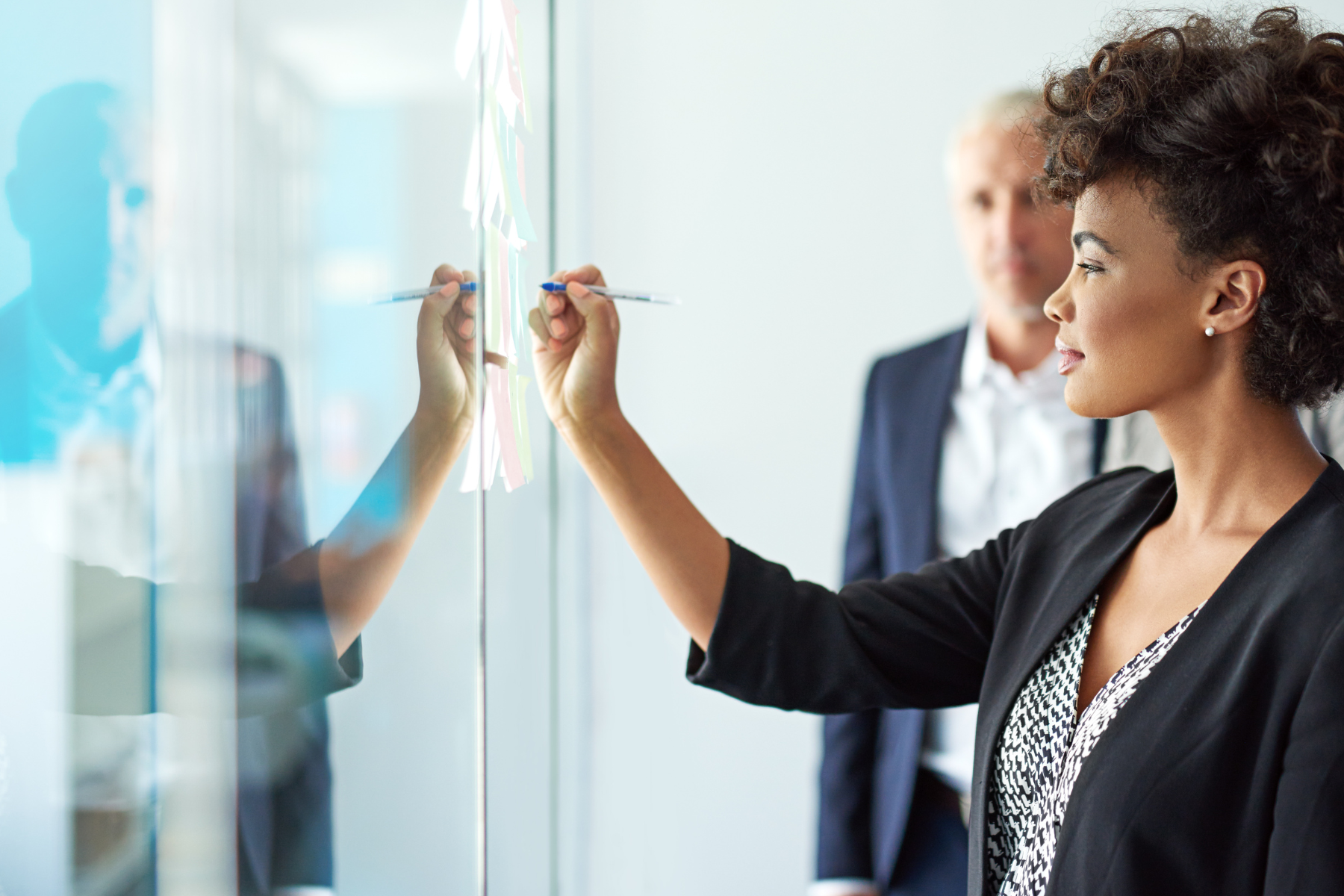 Businesswoman writing on glass board with colleague in the background.