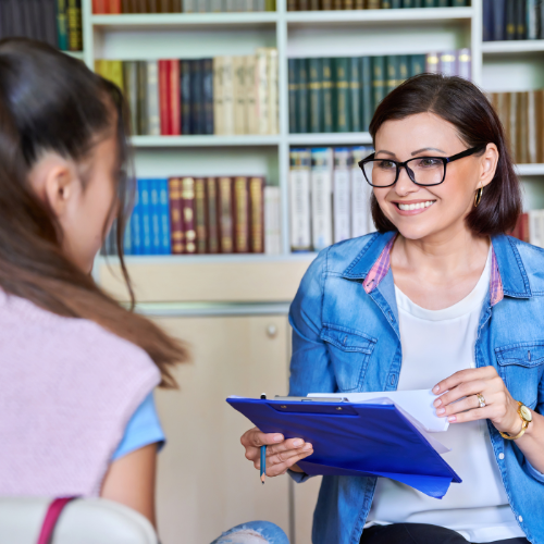 Woman holding clipboard talking to young girl in library setting