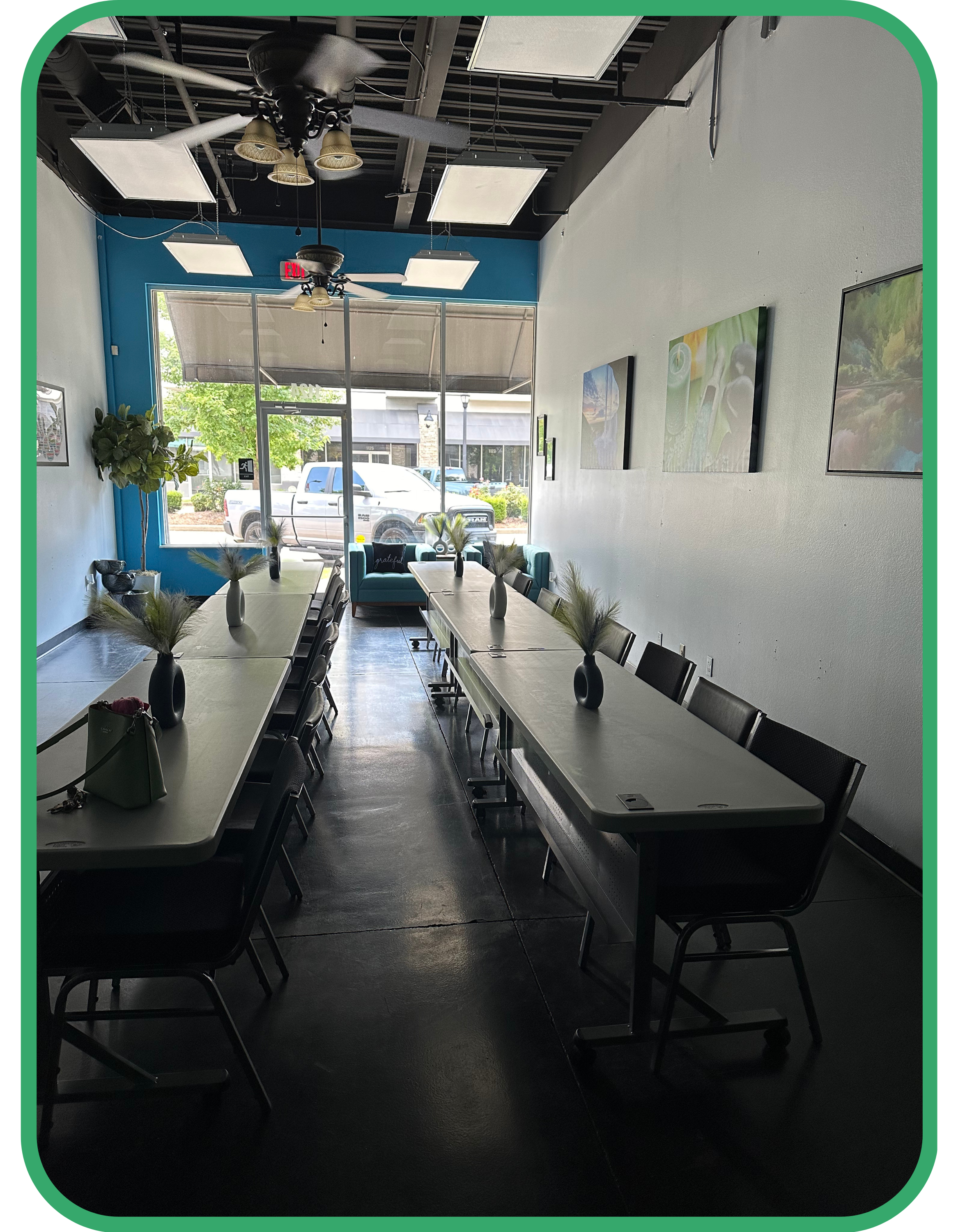 Interior of a modern cafe with long tables, chairs, potted plants, ceiling fans, and artwork on the walls.
