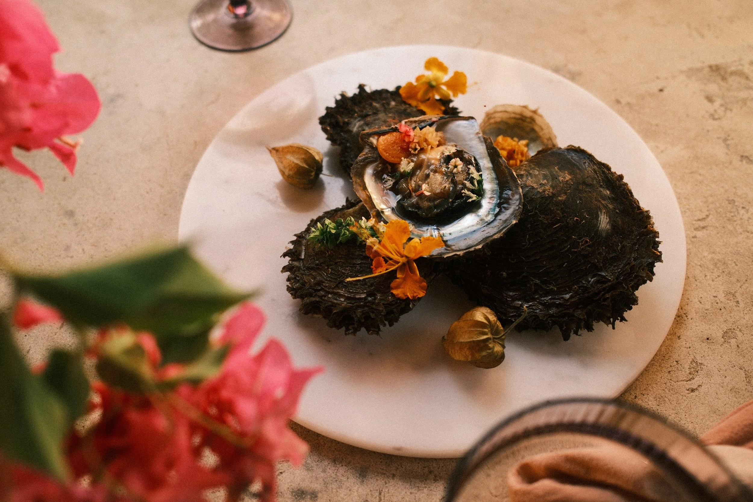 A white plate with a cooked sea creature, garnished with flowers and herbs, surrounded by garlic cloves, on a beige table.