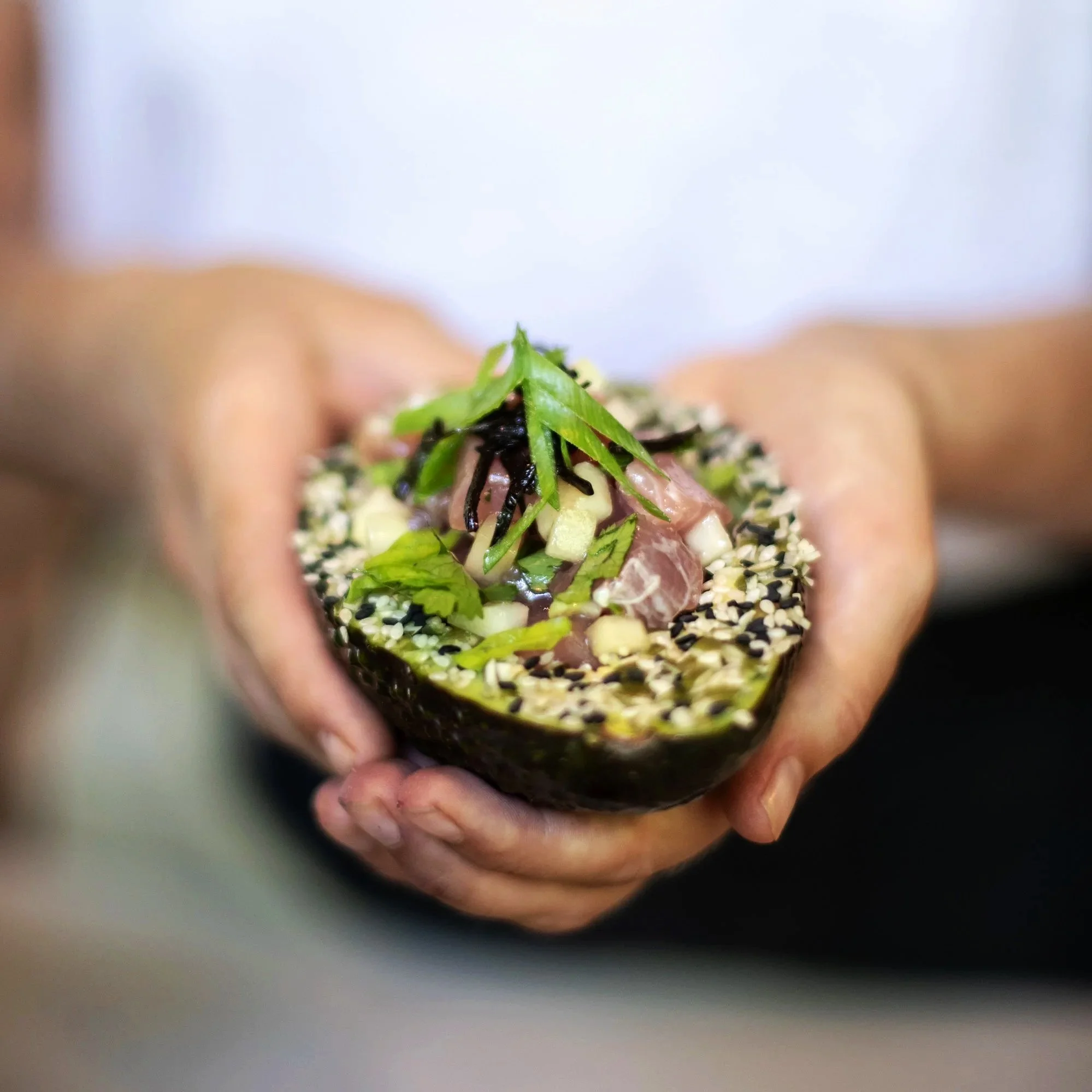 Close-up of a hand holding a halved avocado filled with diced raw fish, topped with chopped green onions, shredded seaweed, and sesame seeds.