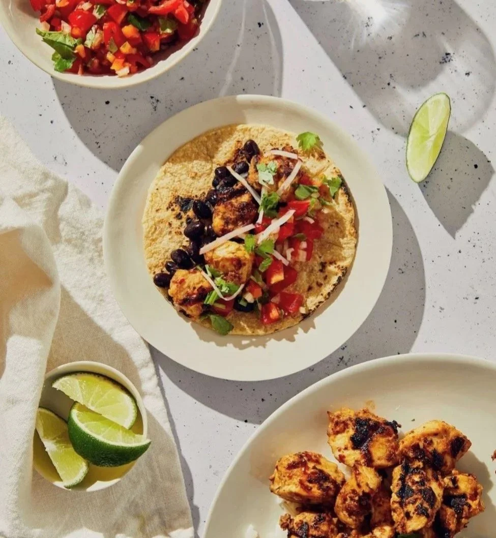 Plate with a taco filled with chicken, black beans, diced tomatoes, shredded cheese, and fresh herbs, on a white speckled surface; a bowl of chopped salsa and a small bowl of lime wedges are also visible.