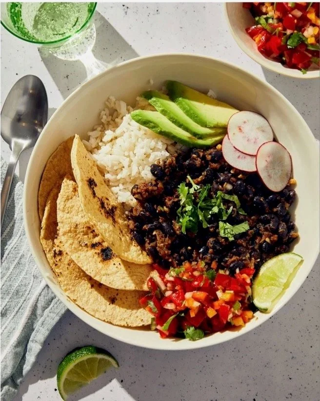 A bowl of Mexican food containing black beans, rice, sliced avocado, radish slices, chopped tomatoes, cilantro, and lime wedges, with a side of tortilla chips. A glass of green beverage and a bowl of salsa are also visible.