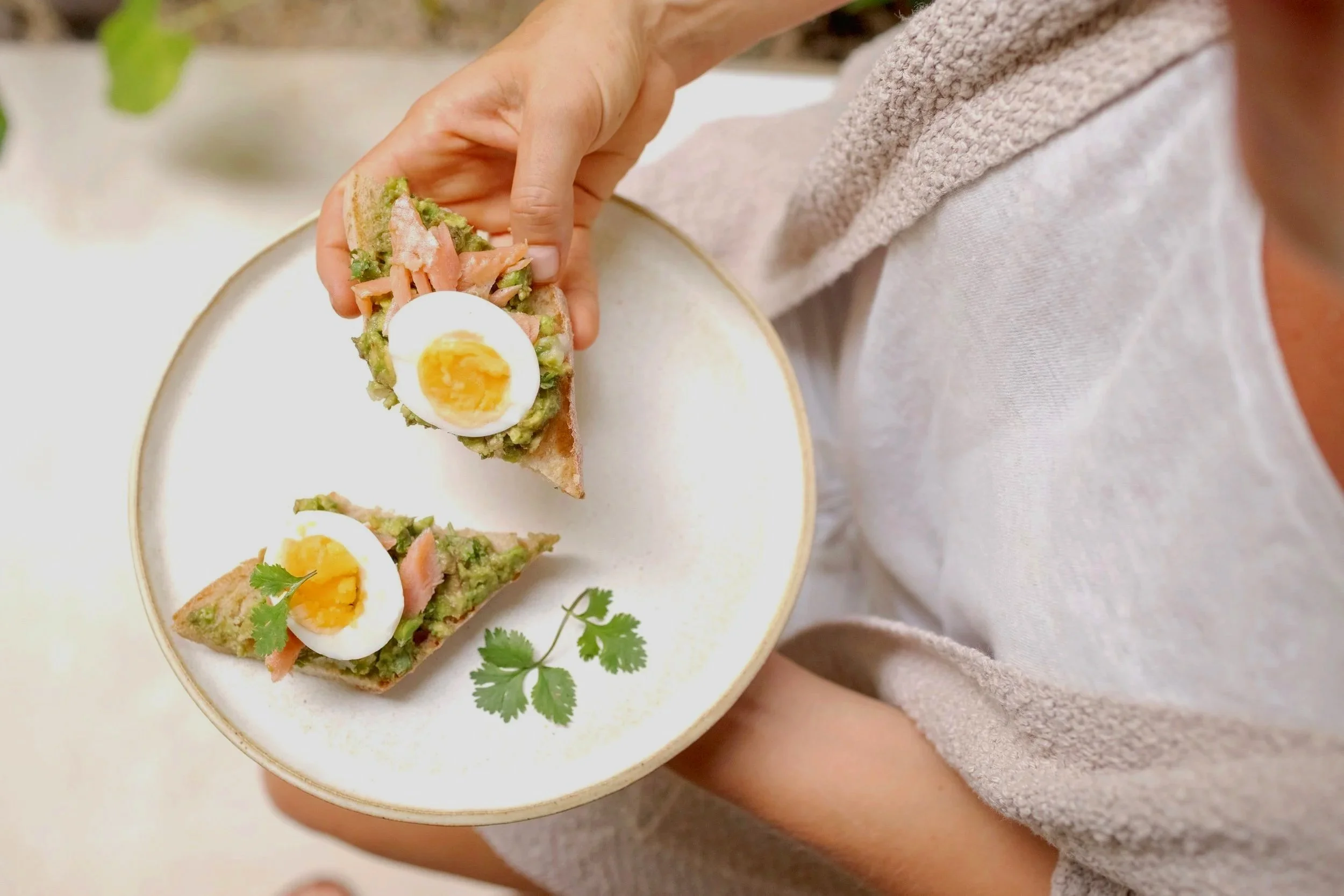 A person holds a white ceramic plate with two slices of avocado toast topped with boiled eggs, salmon, and cilantro.
