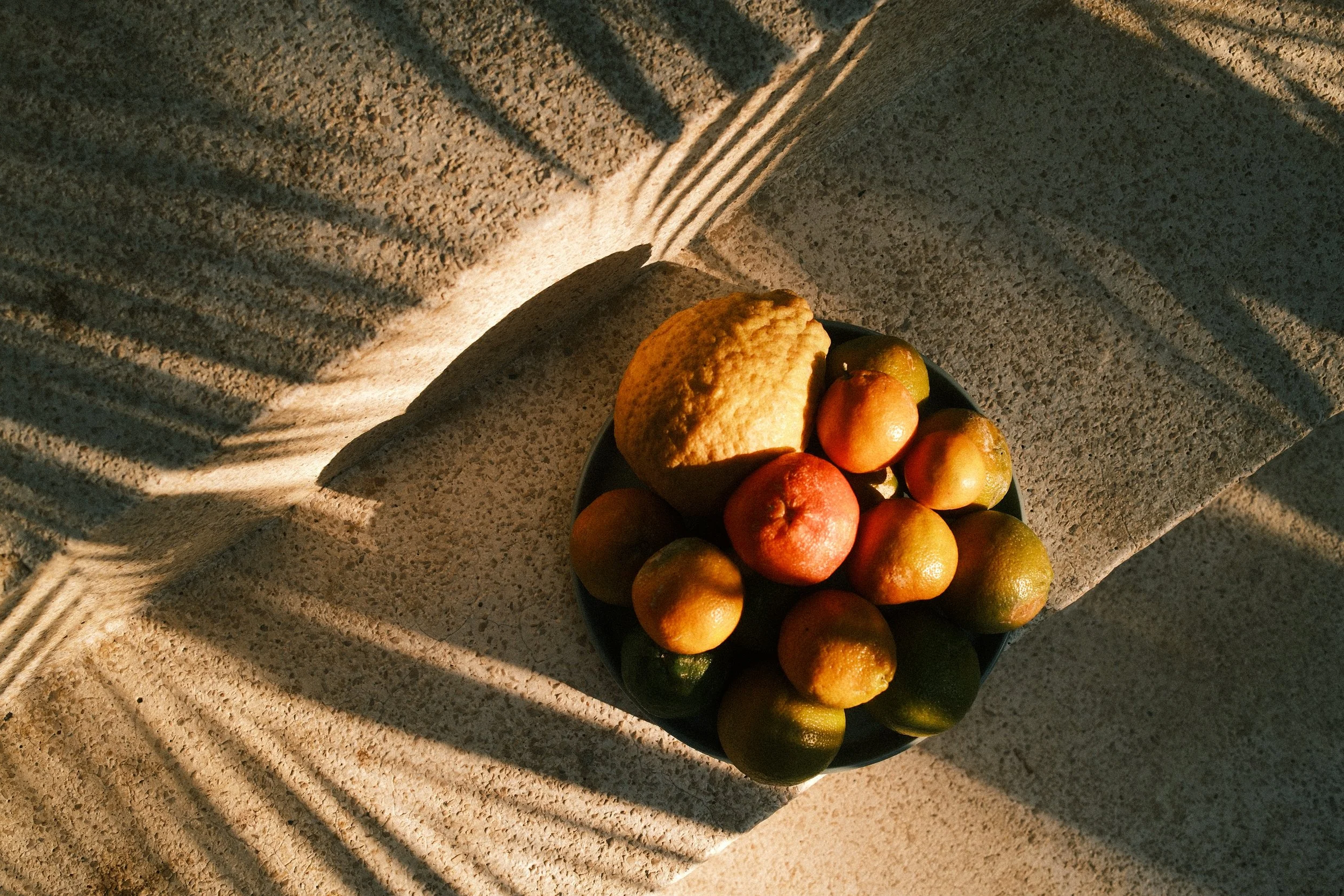 A bowl of assorted citrus fruits, including a lemon, a red apple, and several green and yellow clementines, sitting on a textured stone surface with shadows cast by nearby slats.