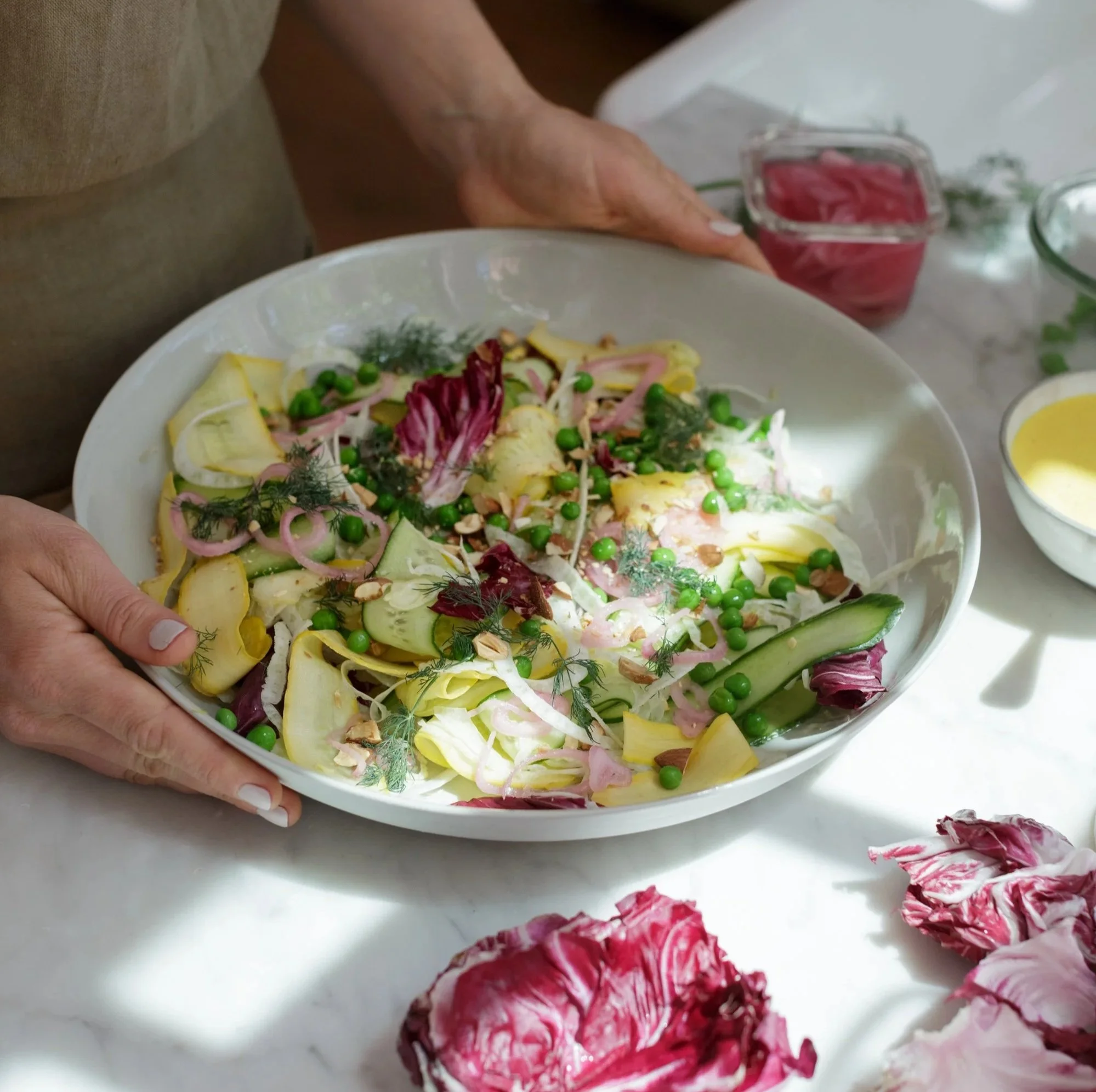 A person holding a white bowl with a colorful vegetable salad including peas, cucumber, radicchio, fennel, and dill, on a white marble surface with radicchio leaves and a small bowl of dressing nearby.