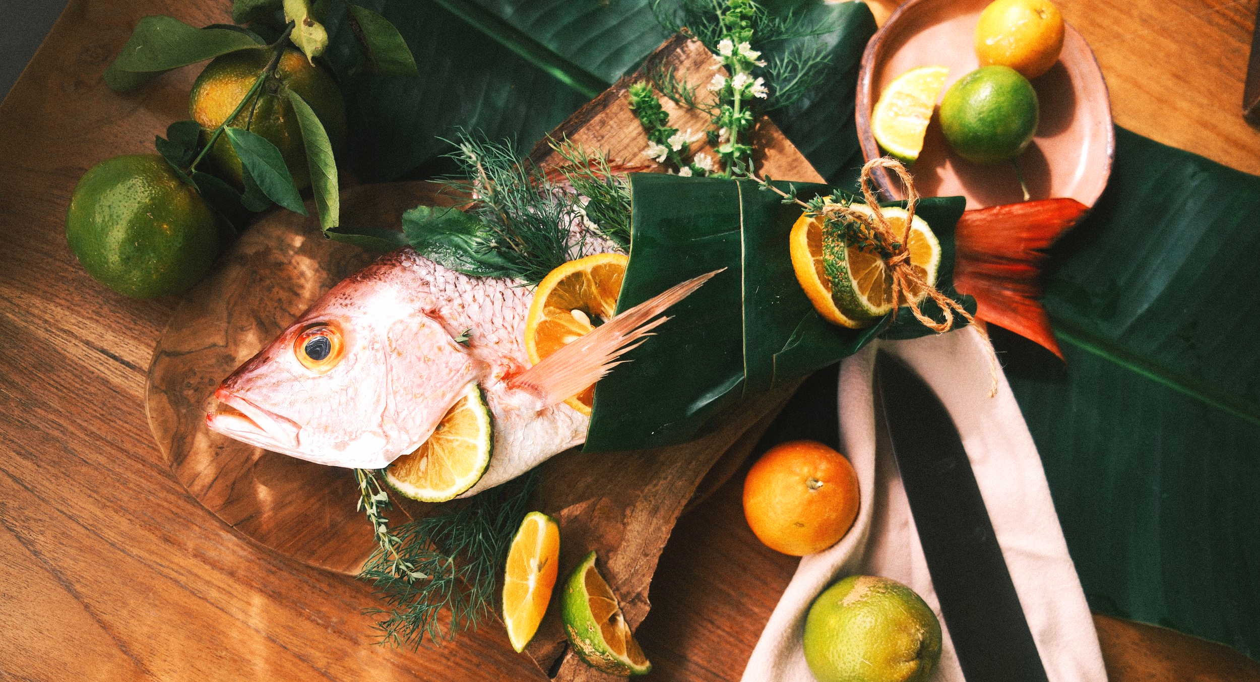 A fish garnished with lemon slices, surrounded by citrus fruits, leaves, and herbs on a wooden surface.