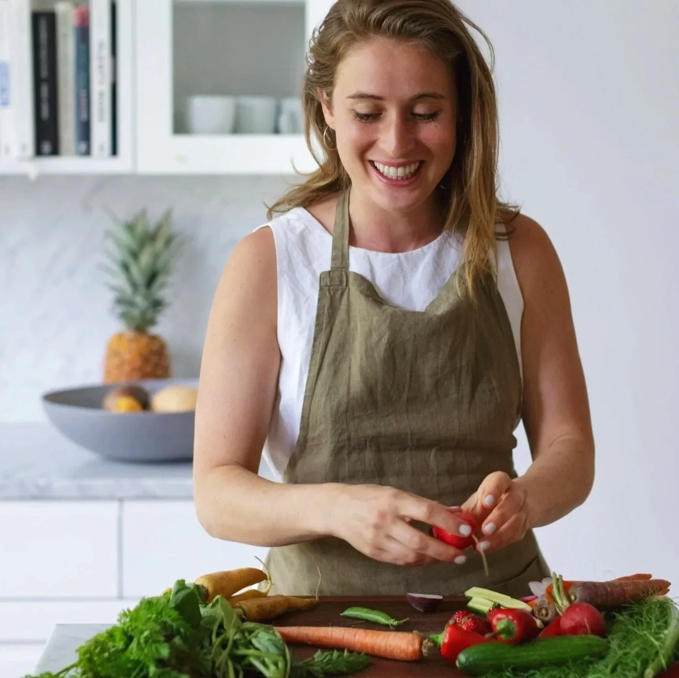 A woman wearing a white sleeveless top and a green apron is peeling a radish in a modern kitchen, with fresh vegetables like carrots, cucumbers, and herbs on the counter in front of her.