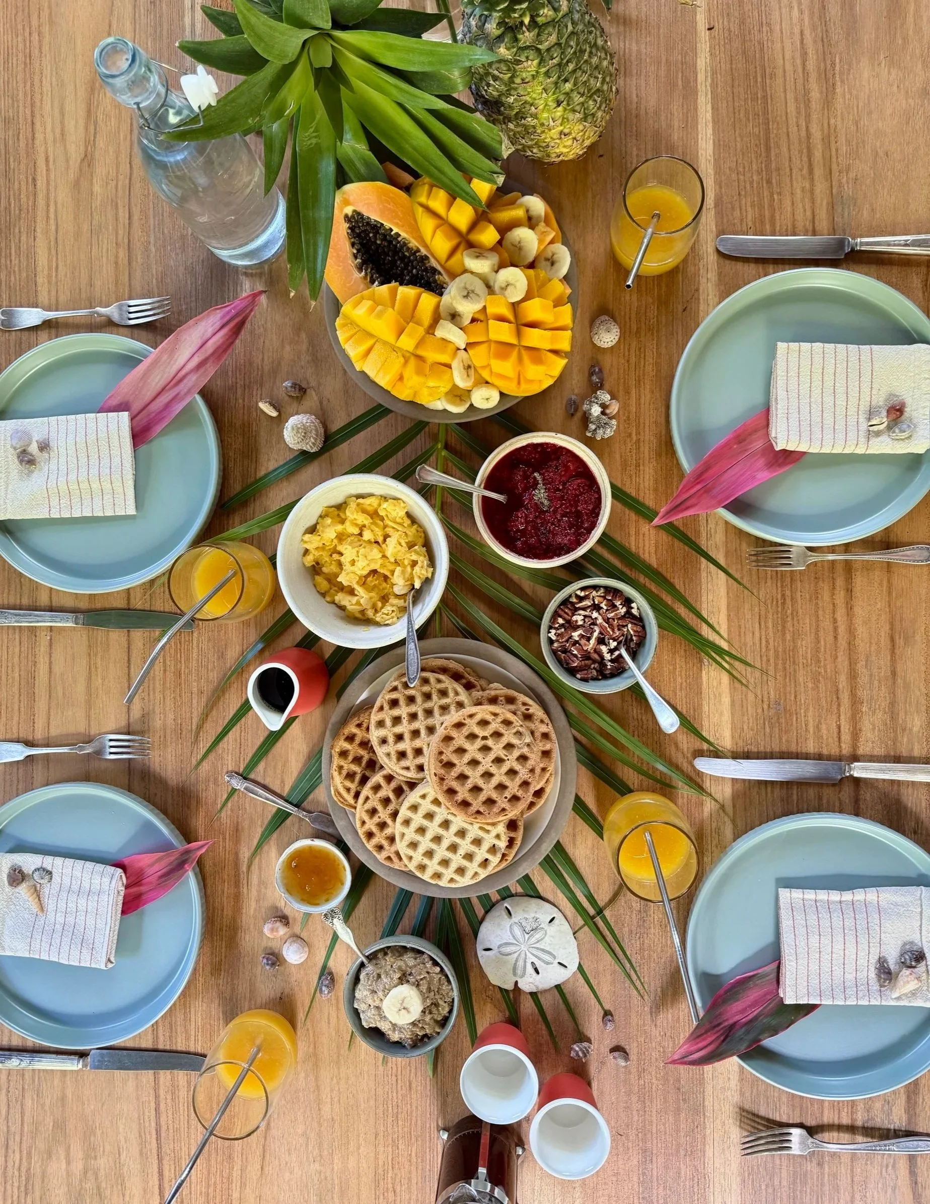 A breakfast table set with four place settings including blue plates, napkins, and cutlery, surrounded by various breakfast foods such as waffles, scrambled eggs, fruit salad with mango, banana, and papaya, pineapple, and sauces, with a centerpiece of tropical fruit and green leaves, and decorative shells and small bowls with toppings and syrup.