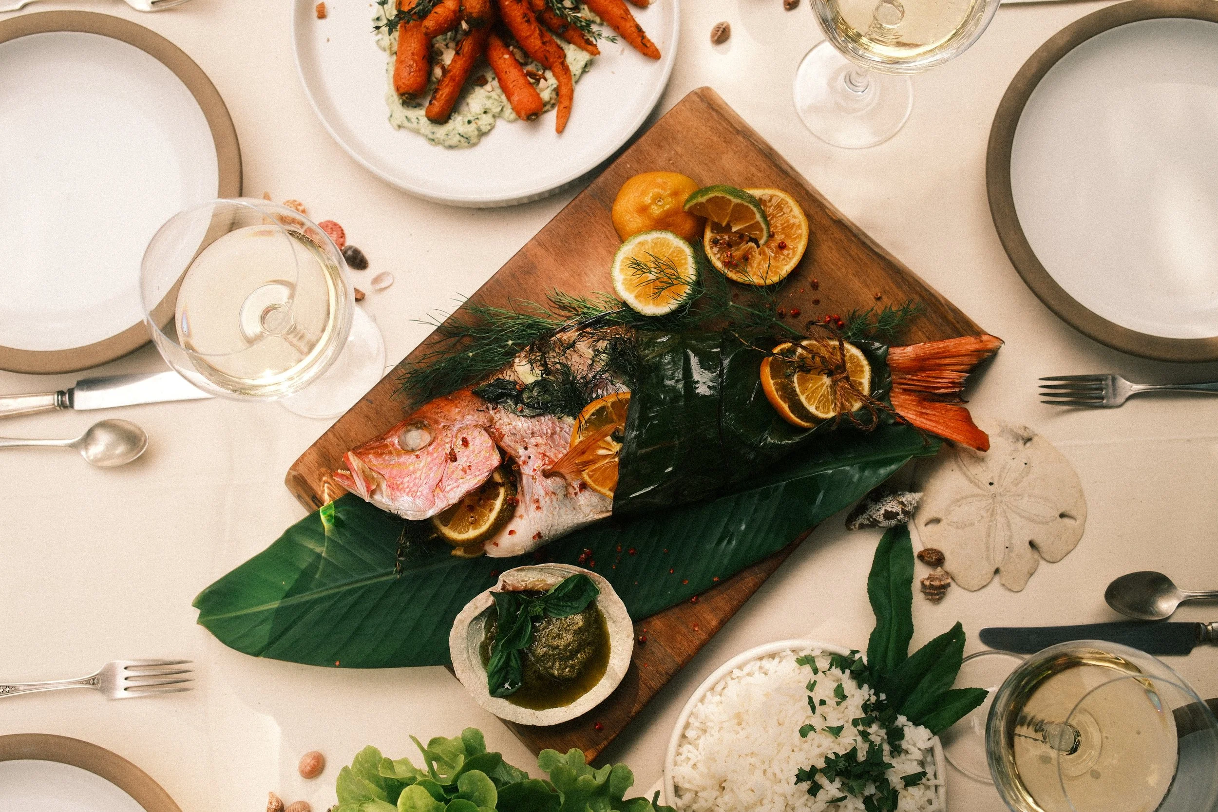 A dining table with a whole fish on a wooden cutting board garnished with lemon slices, herbs, and leafy greens; accompanied by rice, lemon and orange slices, a glass of white wine, and other plates and utensils.