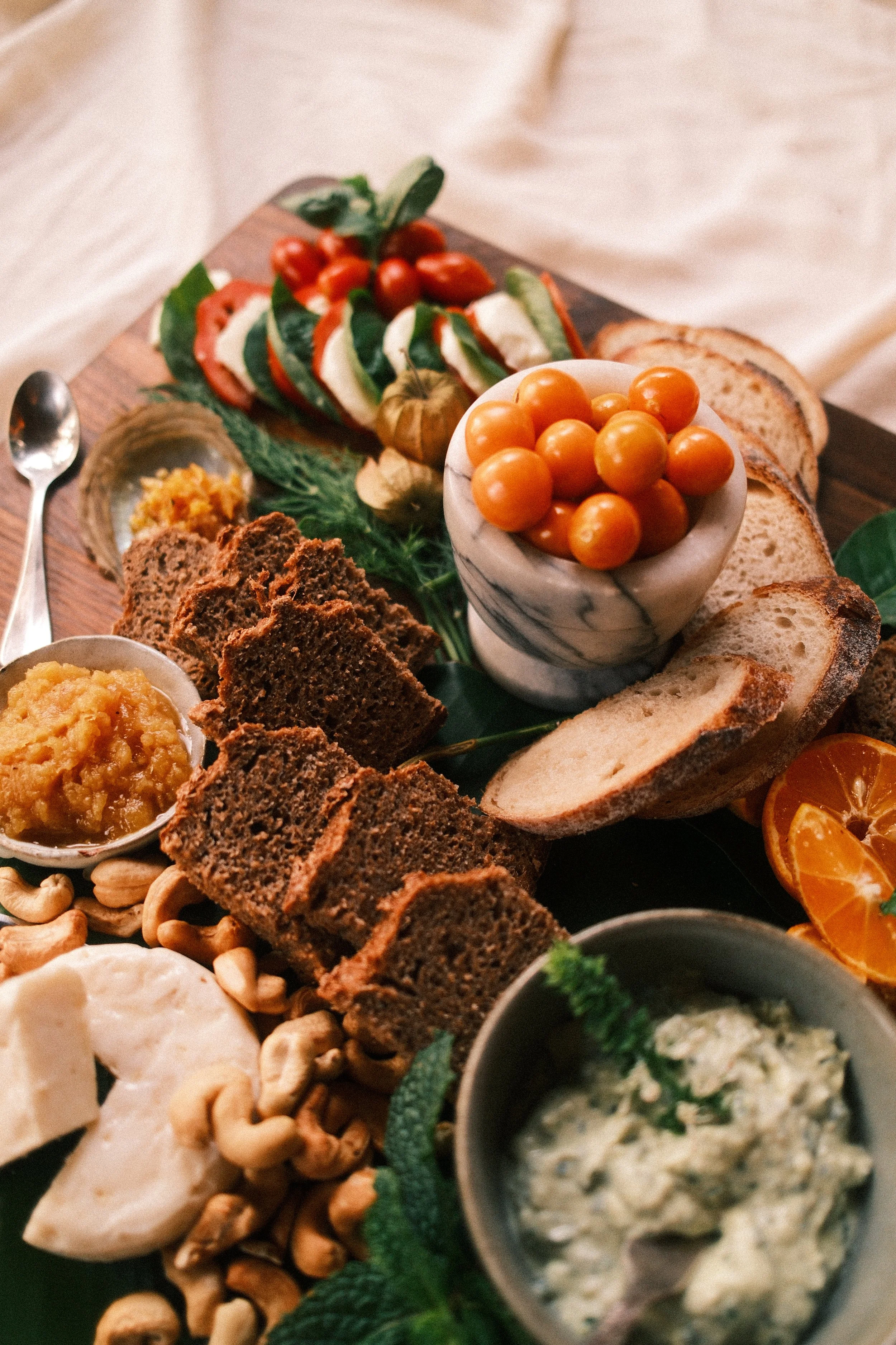 A platters of bread, cherry tomatoes, cheese, nuts, and dips on a wooden board.