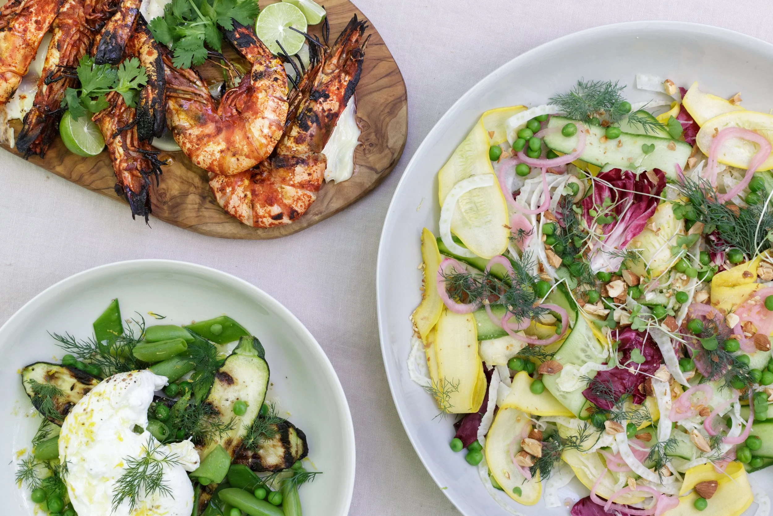 A table with a seafood platter of grilled shrimp and lobster, a bowl of grilled vegetables with a poached egg, and a large bowl of mixed salad with various greens, peas, cucumbers, onions, and nuts.