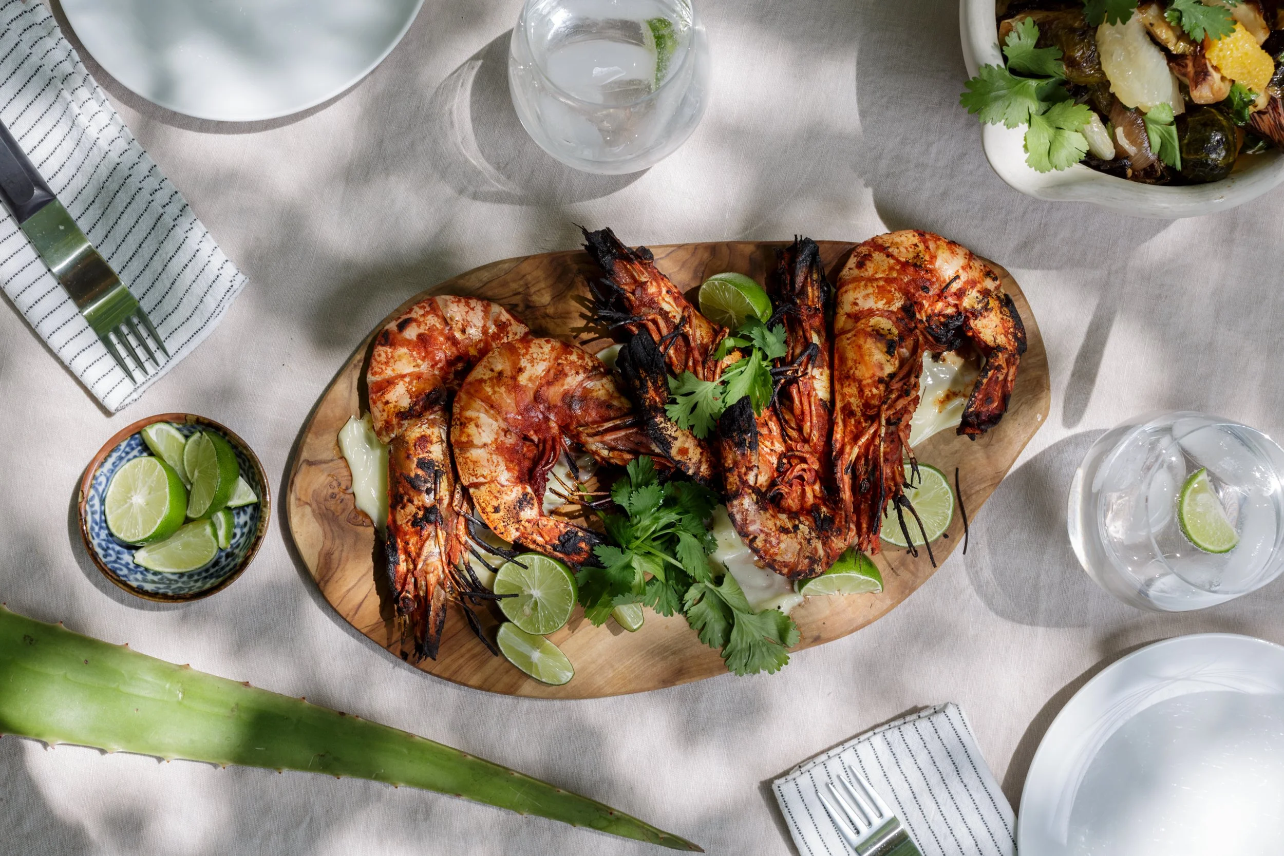 Grilled shrimp served on a wooden platter with lime wedges and fresh cilantro, surrounded by plates, glasses, and utensils on a table with a beige tablecloth.