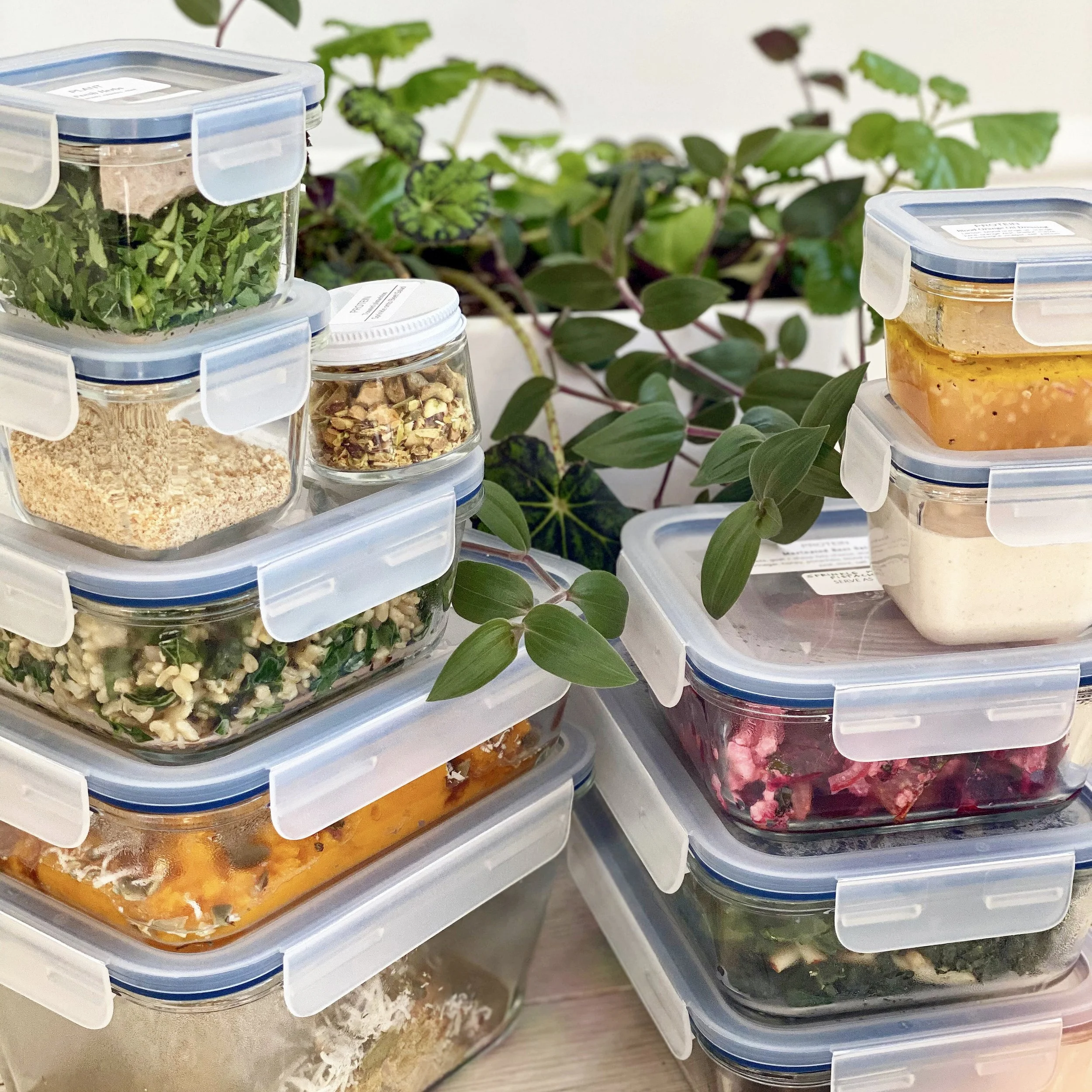 Stacked glass food containers with various salads and dishes, placed on a table near a green leafy plant.