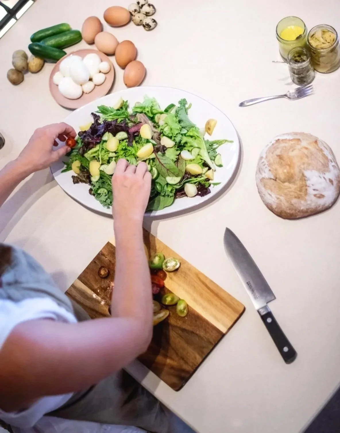 Person chopping cherry tomatoes on a wood cutting board with a knife at a kitchen counter, with a large bowl of salad containing greens, cucumbers, and potatoes, and various eggs and ingredients nearby.