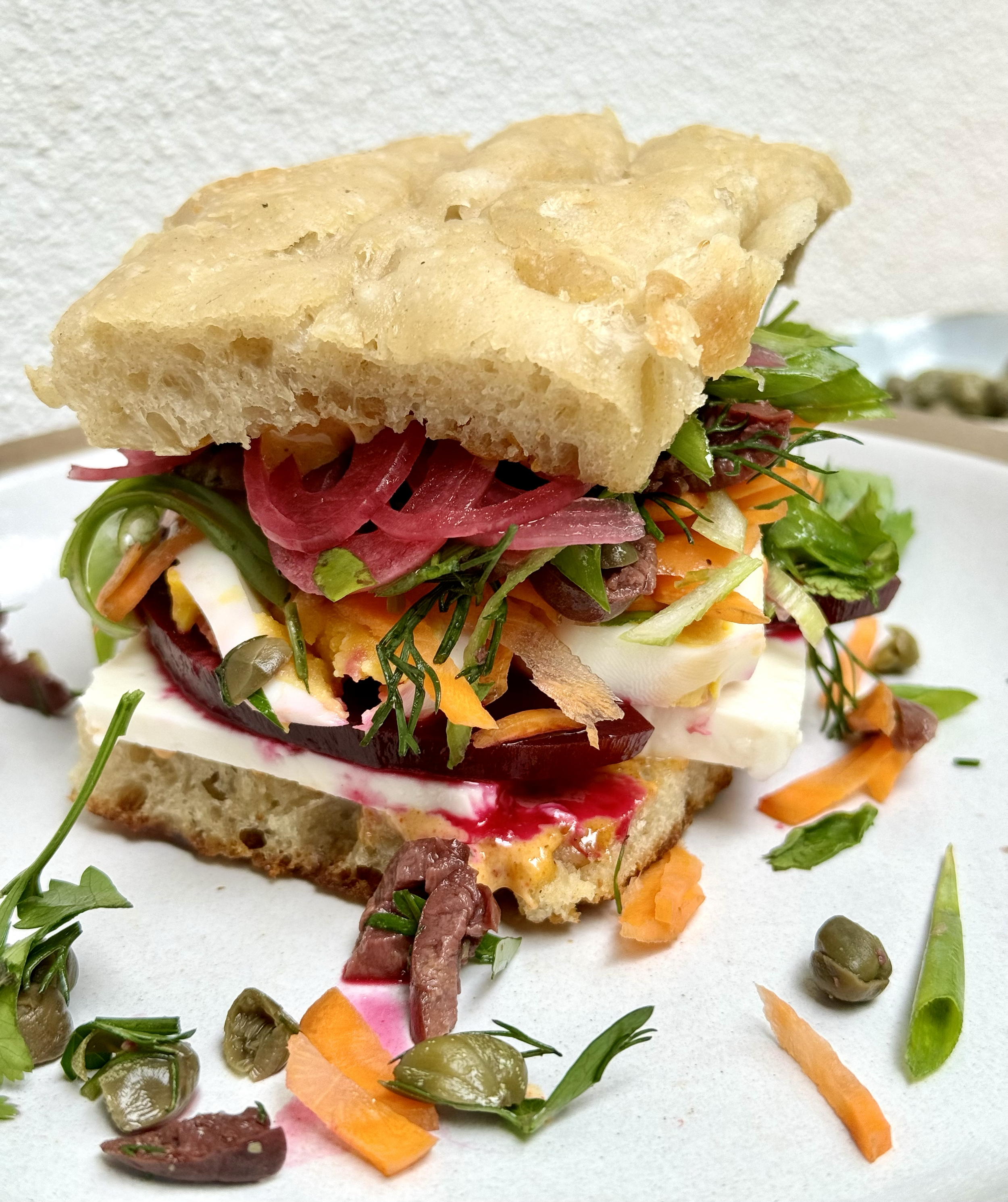 Close-up of a sandwich with various colorful vegetables, sliced meats, cheese, and a piece of bread on top, with some salad ingredients scattered around.