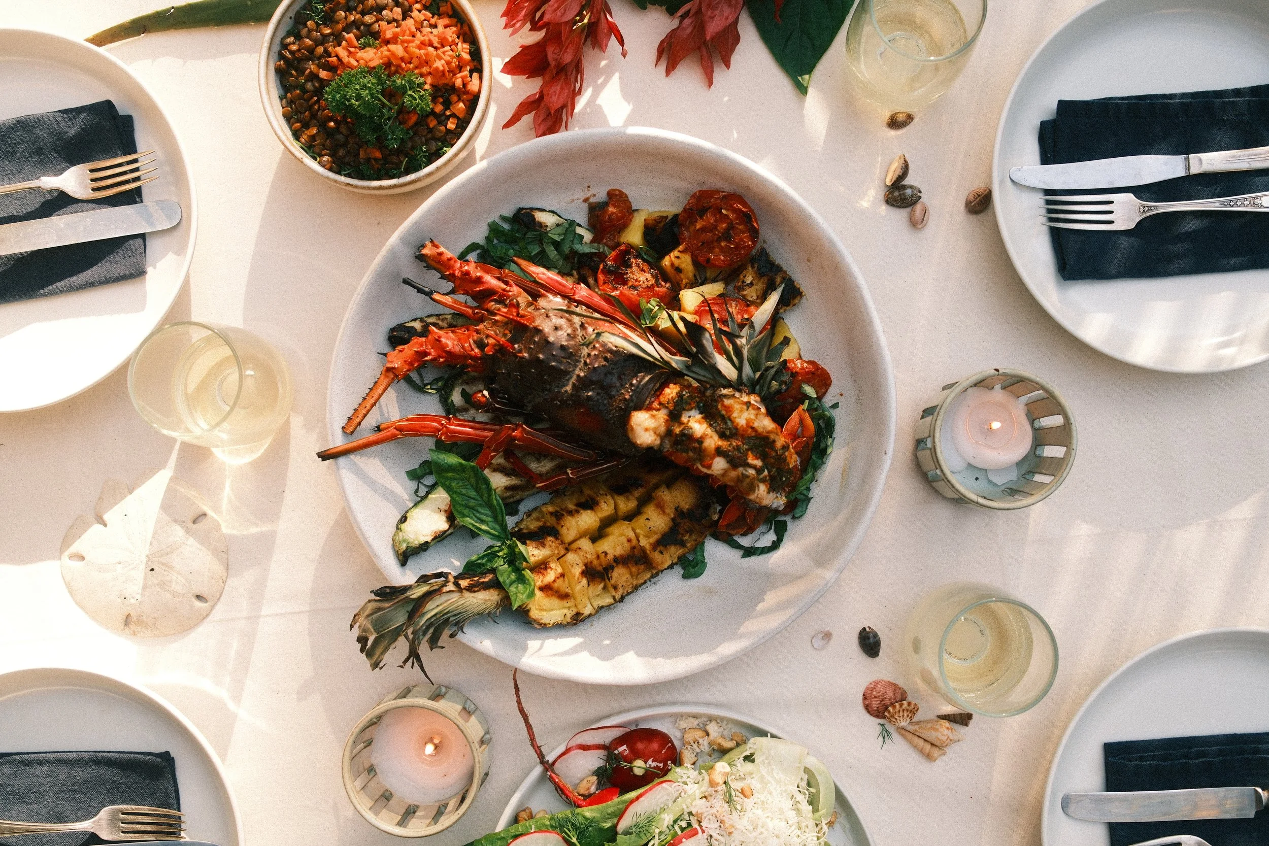 Seafood dish with lobster and grilled vegetables on a white plate, surrounded by salad, candles, and drinks on a table set for a meal.