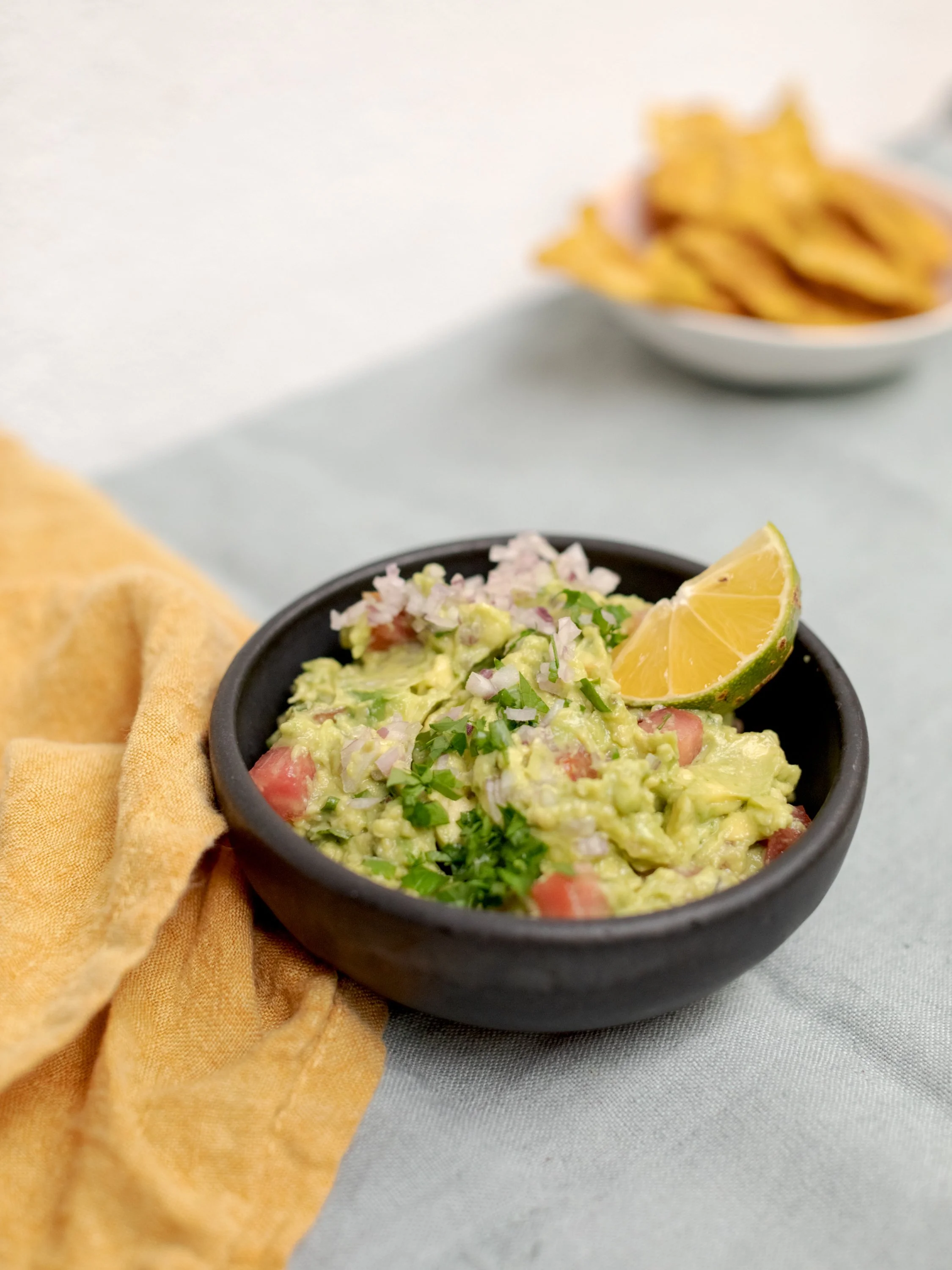 A bowl of guacamole garnished with chopped onions, tomatoes, cilantro, and a lime wedge, with a bowl of tortilla chips in the background.