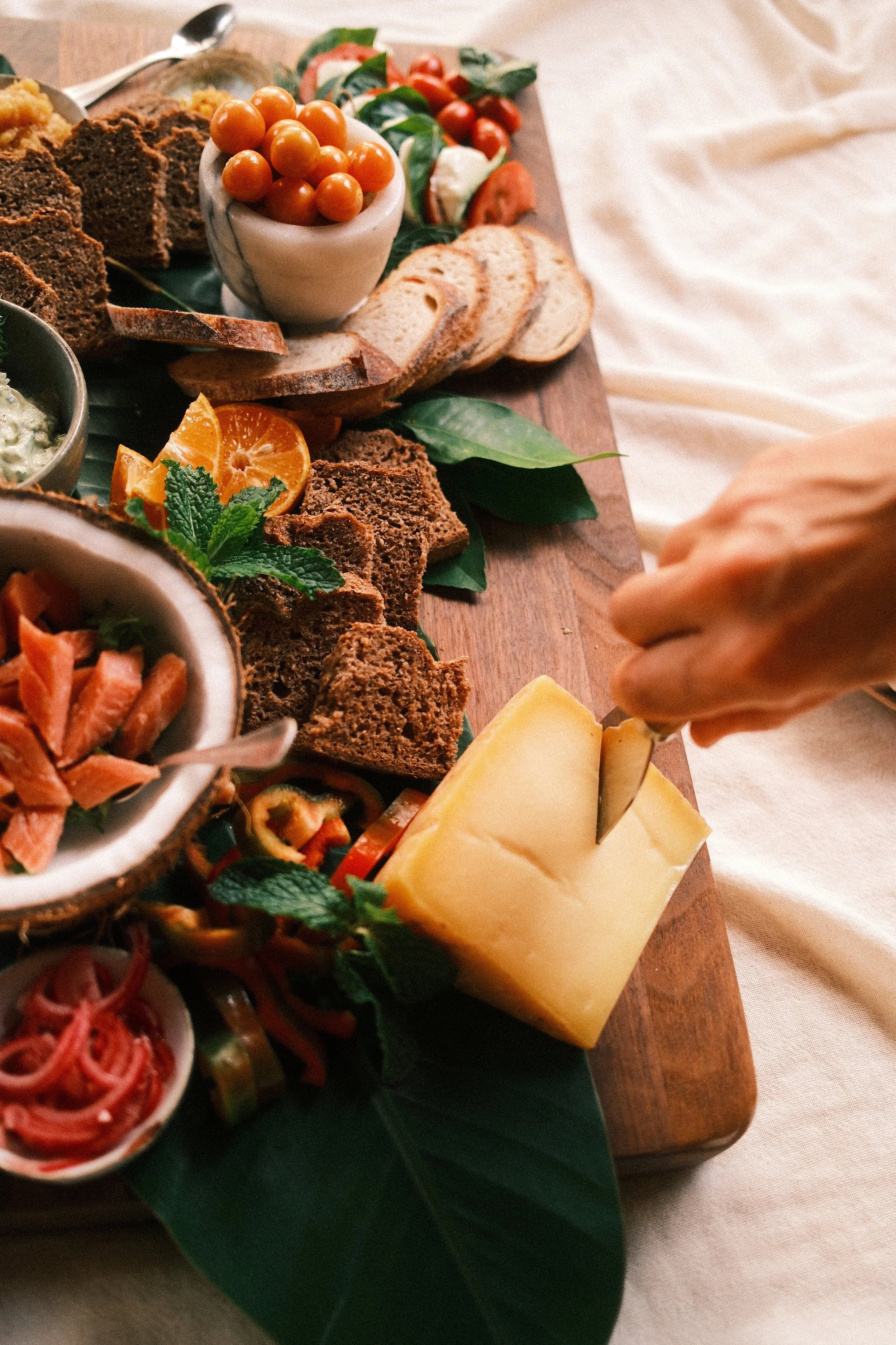 A cheese board with sliced bread, cherry tomatoes, assorted cheeses, and various spreads and garnishes, served on a wooden cutting board.