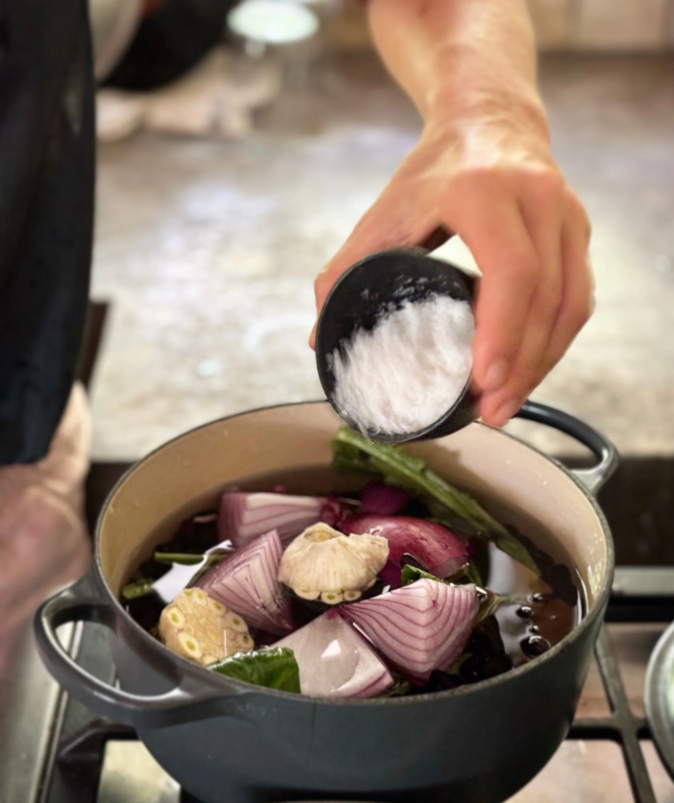 Person adding salt to a pot of chopped vegetables, including onions and garlic, on a stovetop.