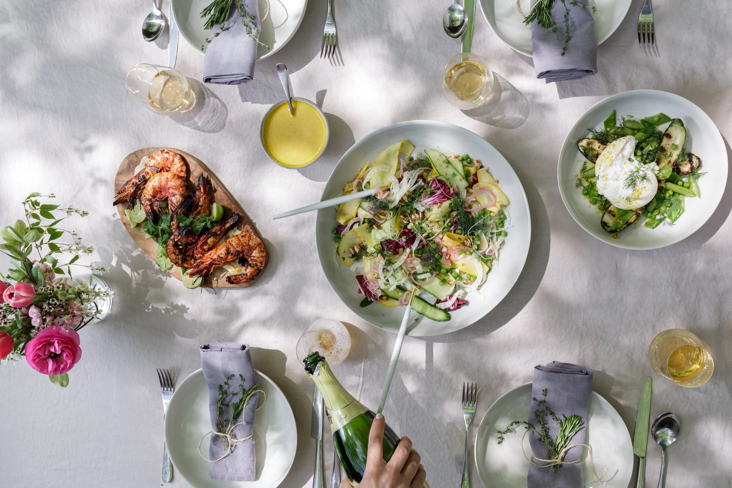 Overhead view of a table set for a meal with a large salad, grilled vegetables, grilled shrimp, drinks, flowers, and place settings with cloth napkins and utensils.