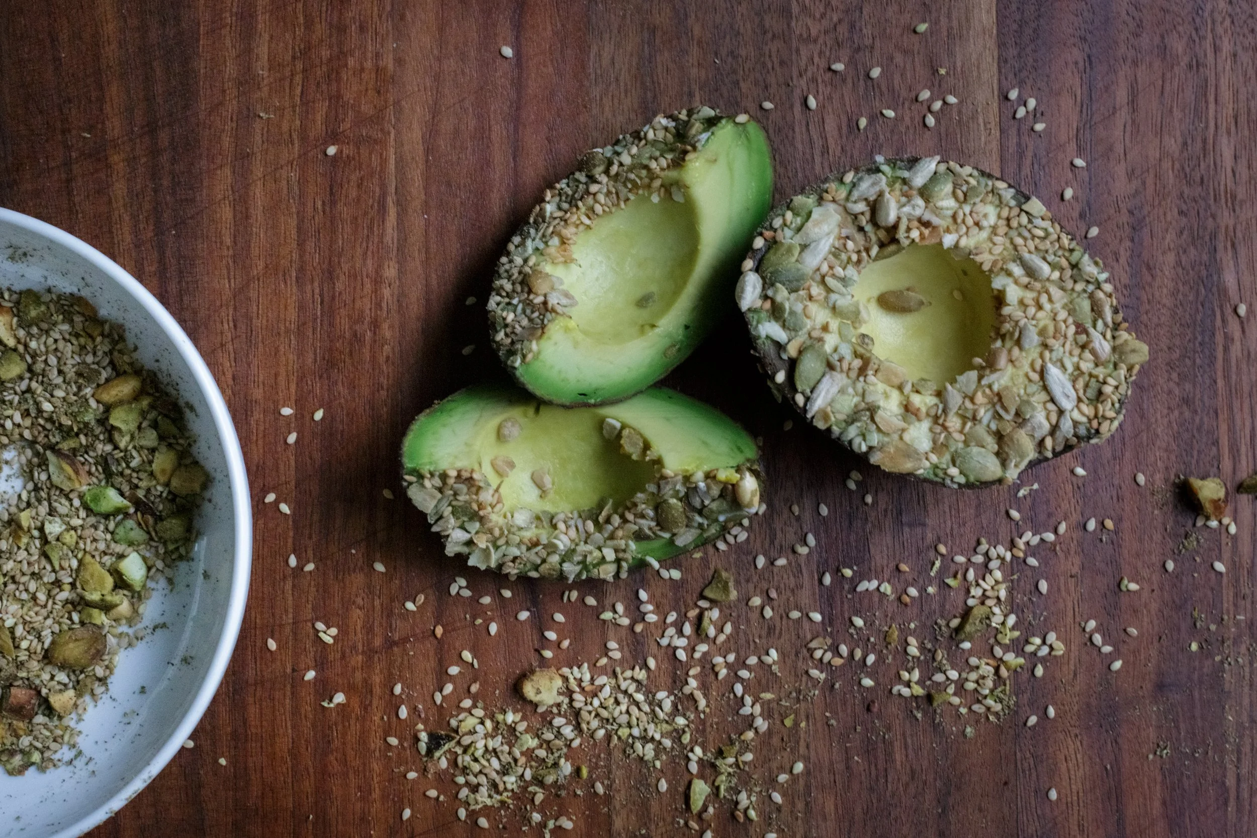 A wooden surface with halved avocado coated in seeds and nuts, and a white bowl filled with similar seed and nut mixture.