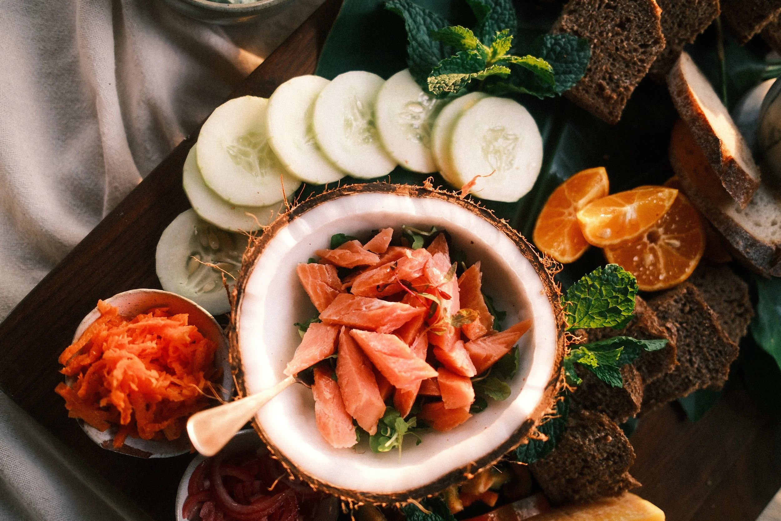 A wooden platter with sliced cucumbers, orange segments, dark bread slices, chopped carrots, and salmon pieces, garnished with mint leaves.