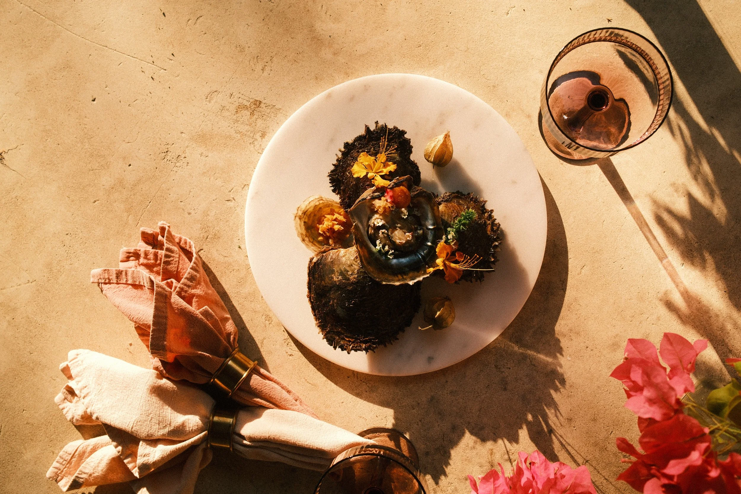 A cooked oyster on a white plate surrounded by decorative flowers, with a pink napkin and a glass of drink on a beige surface.
