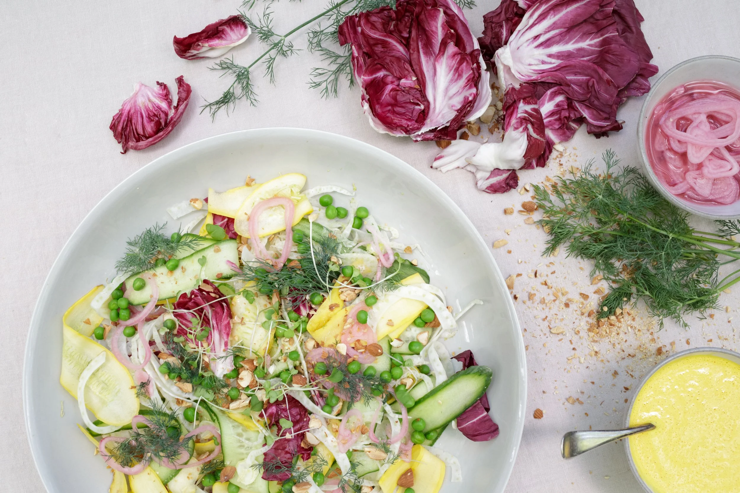 A fresh vegetable salad with cucumbers, radicchio, green peas, fennel, and herbs served on a plate, with scattered radicchio leaves, herbs, crushed nuts, and a bowl of pink pickled onions nearby on a light-colored table.