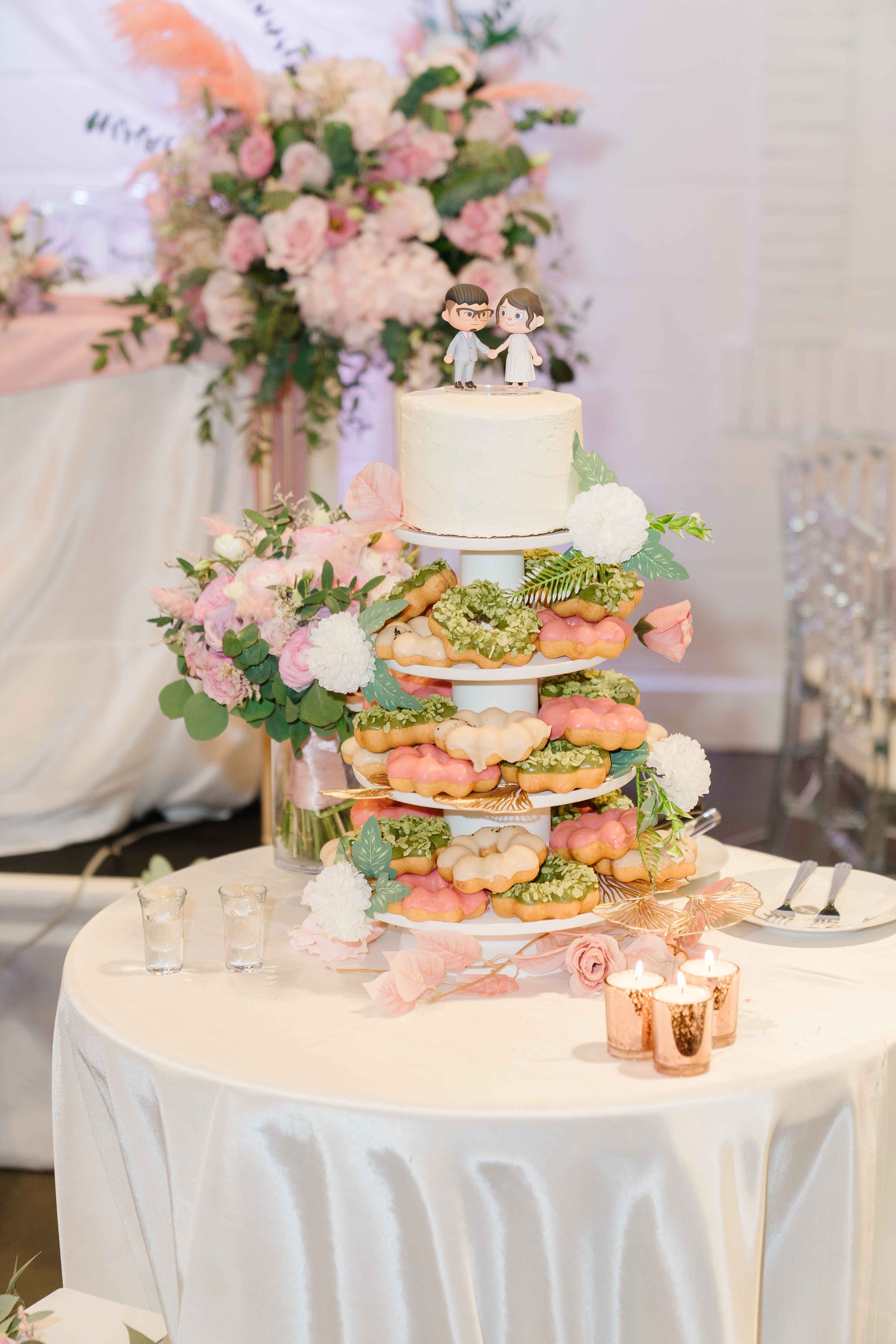 A wedding cake with a miniature bride and groom topper, decorated with pink, green, and white flowers, surrounded by colorful cookies on a wedding reception table with pink candles and floral arrangements.