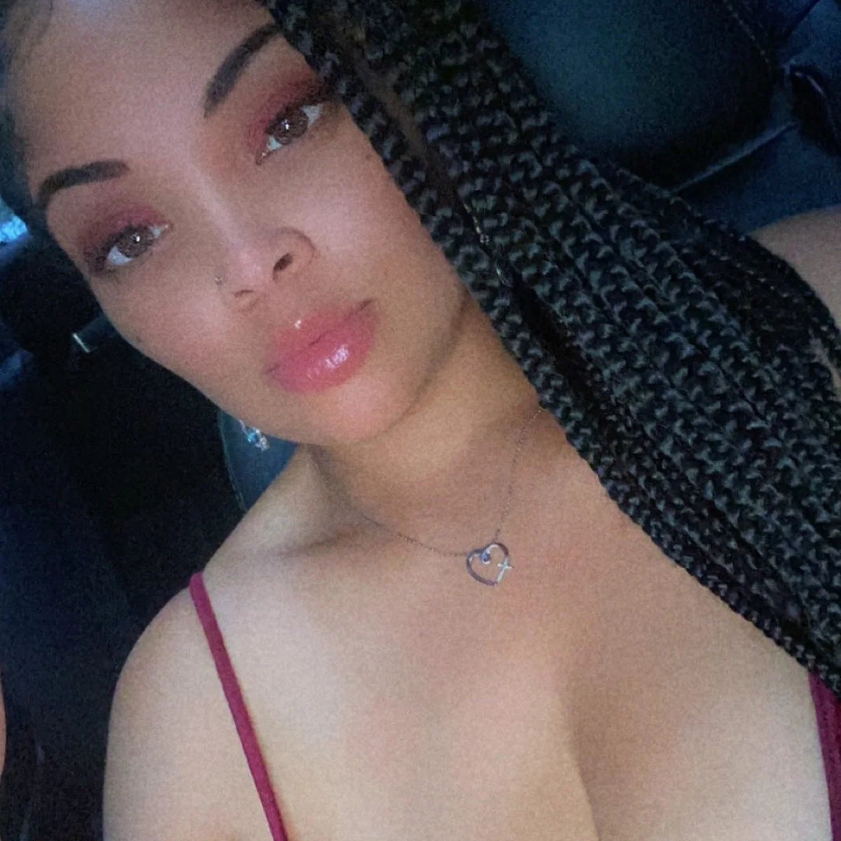 A woman with long braided hair and light makeup, wearing a pink top and a heart-shaped necklace, sitting inside a vehicle.