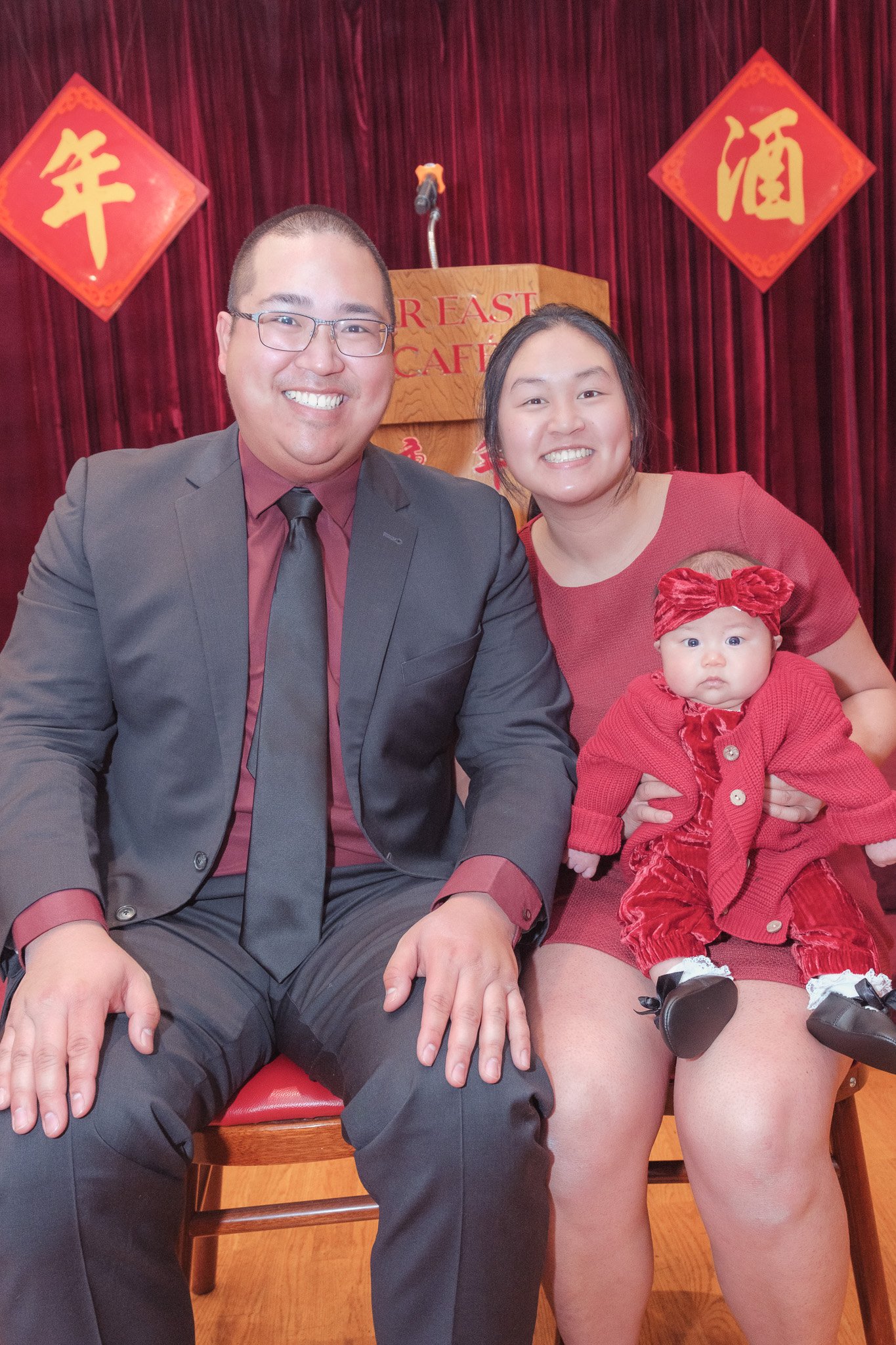 Family sitting together at a celebration, with a man in a suit, a woman holding a baby girl dressed in red, and Chinese New Year decorations in the background.
