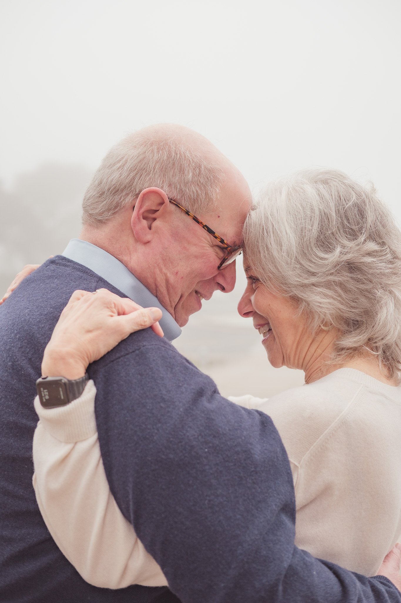 An elderly man and woman embrace, touching foreheads together and smiling, with a foggy outdoor background.