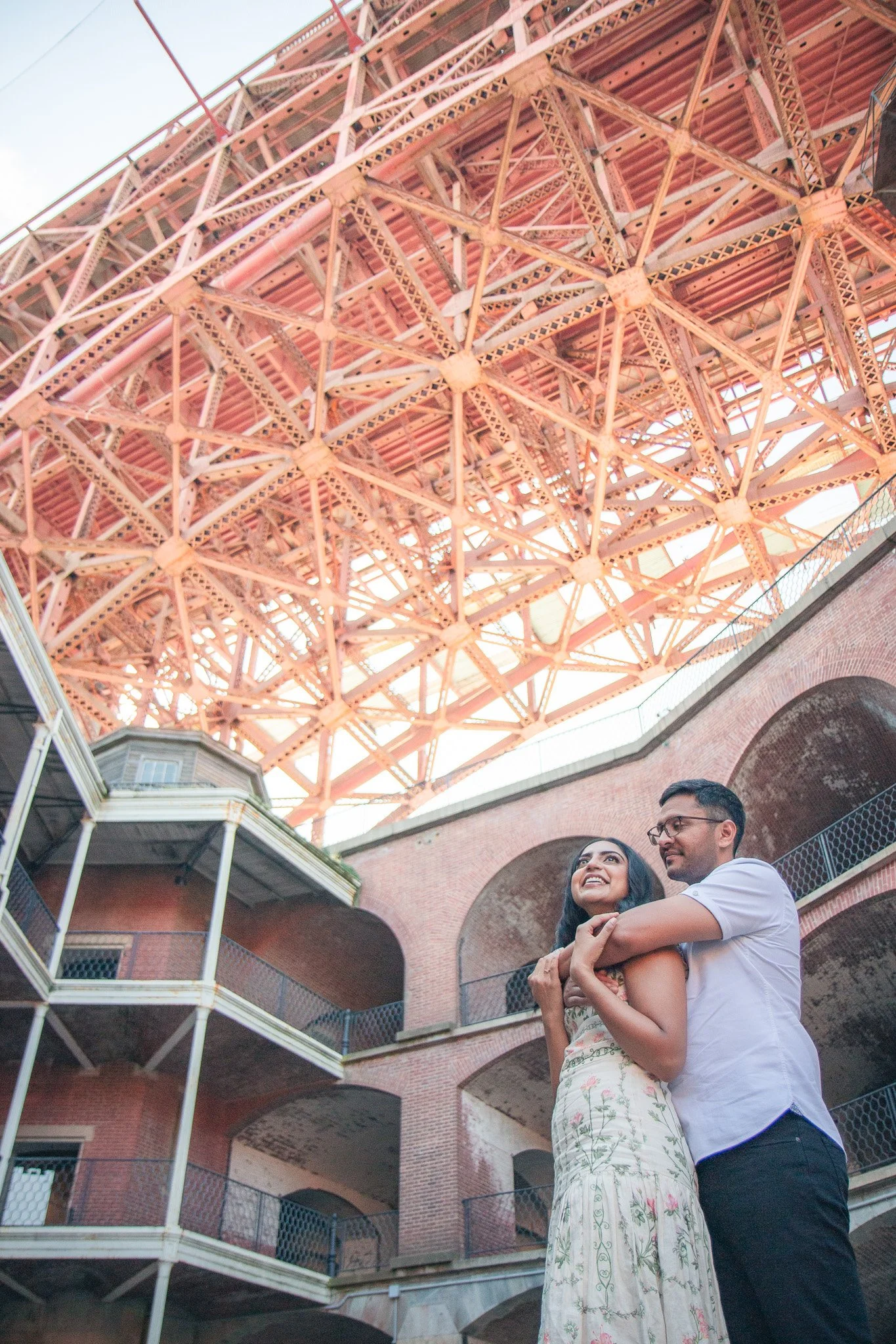 A couple embraces under a large, intricate, pink steel bridge structure with a brick building in the background.