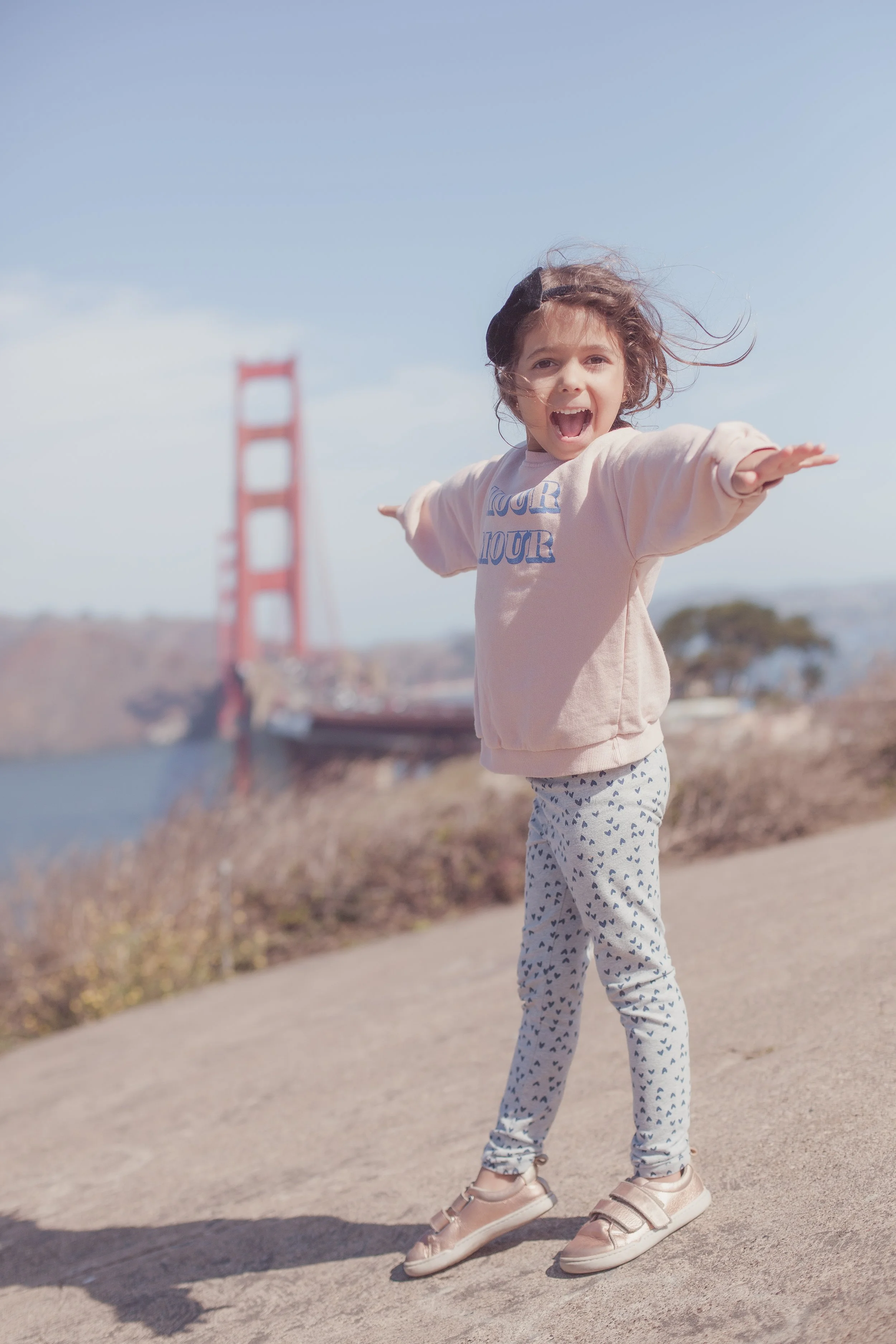 A young girl with curly brown hair, wearing a light pink sweatshirt with blue text, patterned pants, and beige sneakers, standing on a sidewalk near the Golden Gate Bridge in San Francisco, with arms outstretched and smiling at the camera.