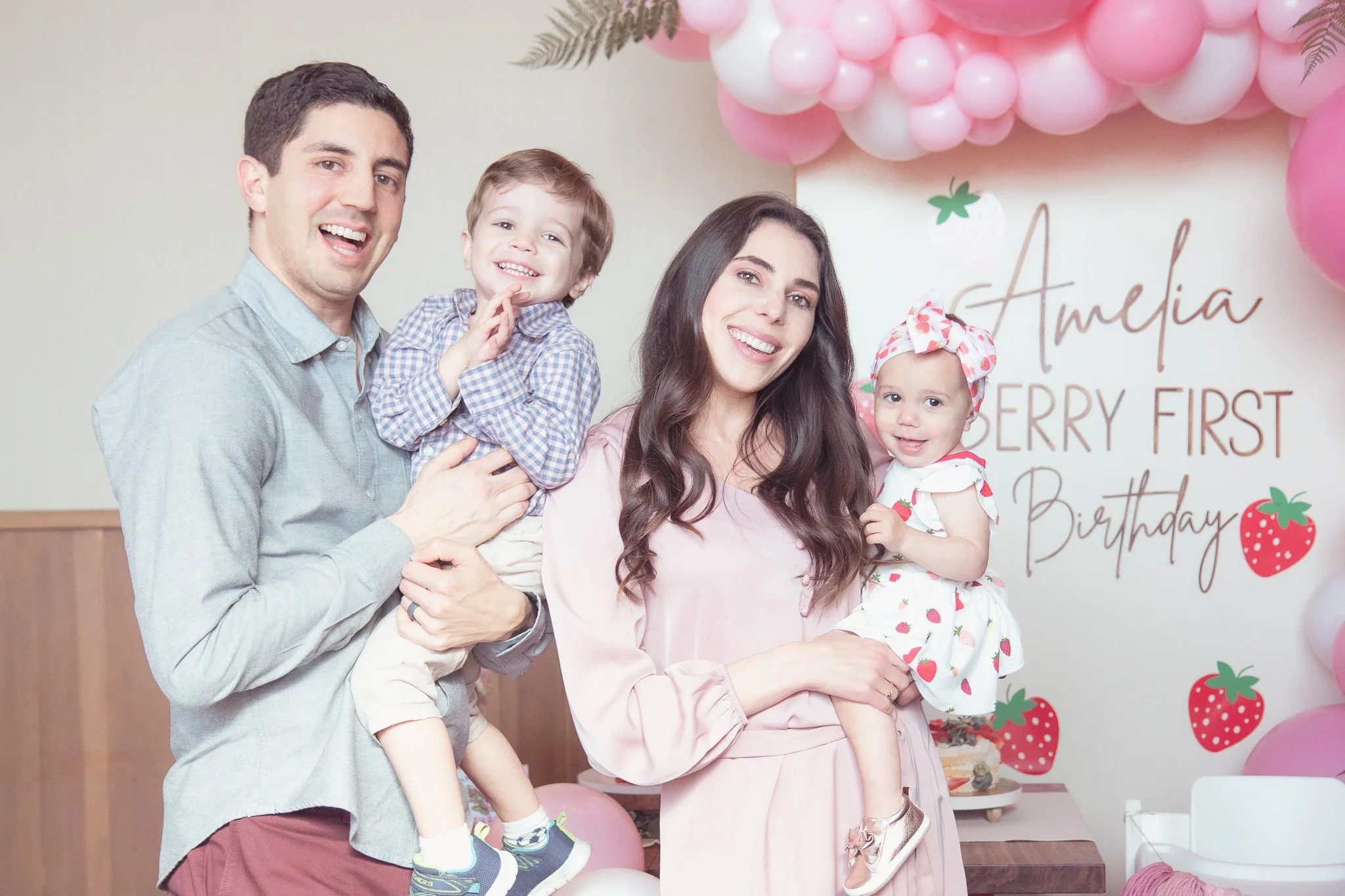 Family celebrating a strawberry-themed birthday party with balloons and a decorated backdrop.