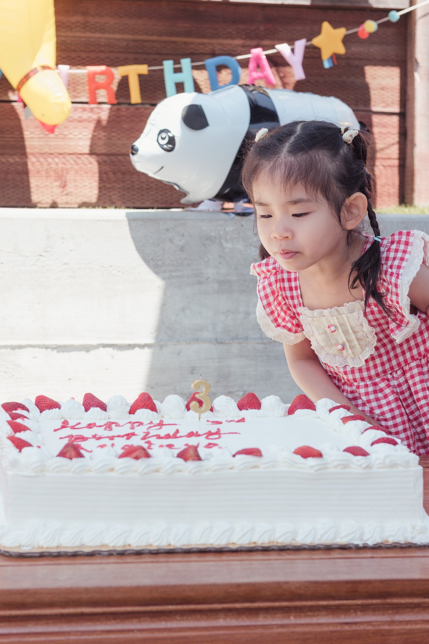 Young girl in gingham dress looking at a large birthday cake decorated with strawberries and whipped cream, with a birthday candle shaped like the number 3. There is a candy-themed birthday banner and a panda-shaped balloon in the background, indicat